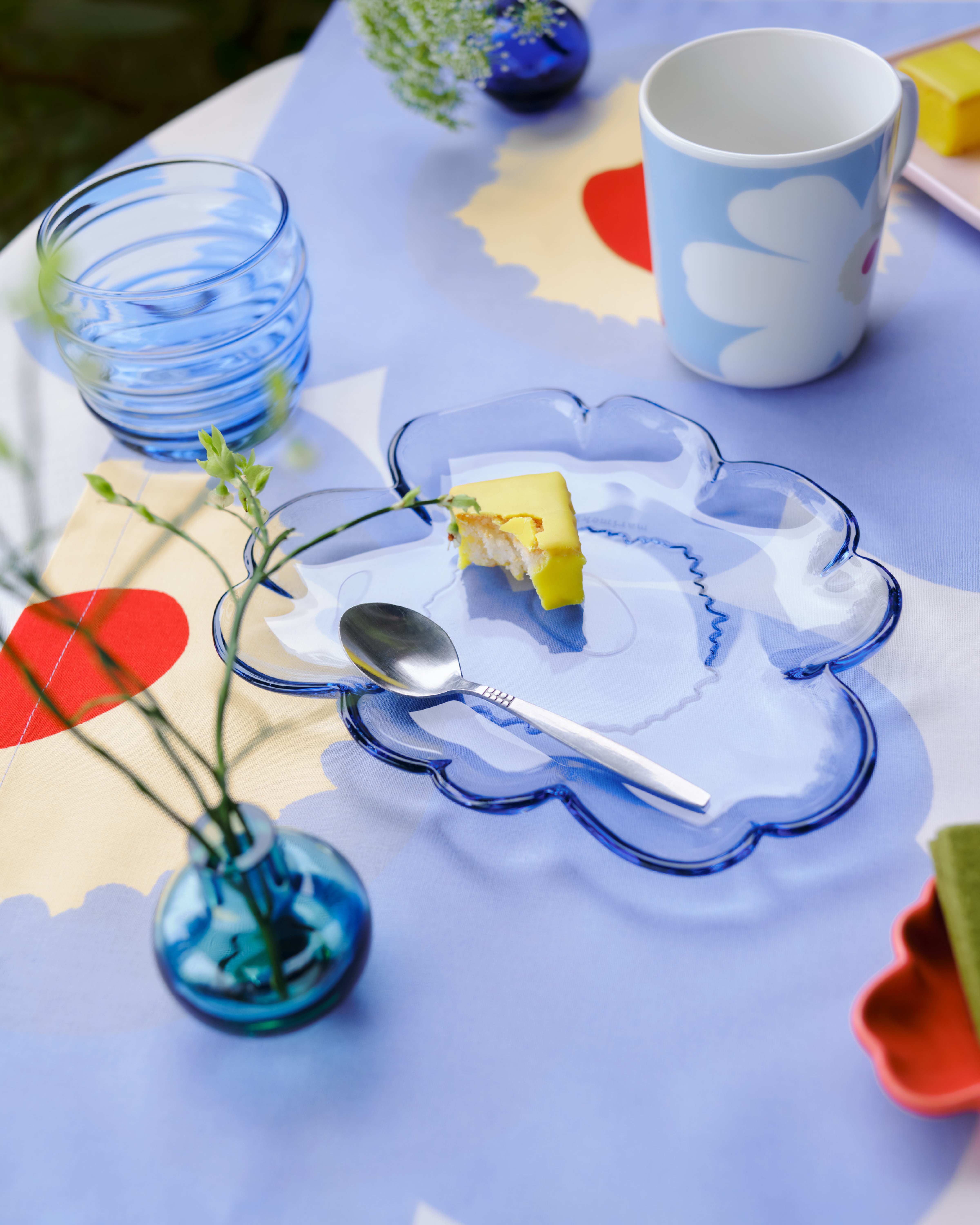 Pöytä ulkona, jossa on siniset Sukat Makkaralla -lasit, sininen Unikko-muotoinen lasilevy ja minimalaiset maljakot Unikko-painatuksella varustetulla pöytäliinalla.