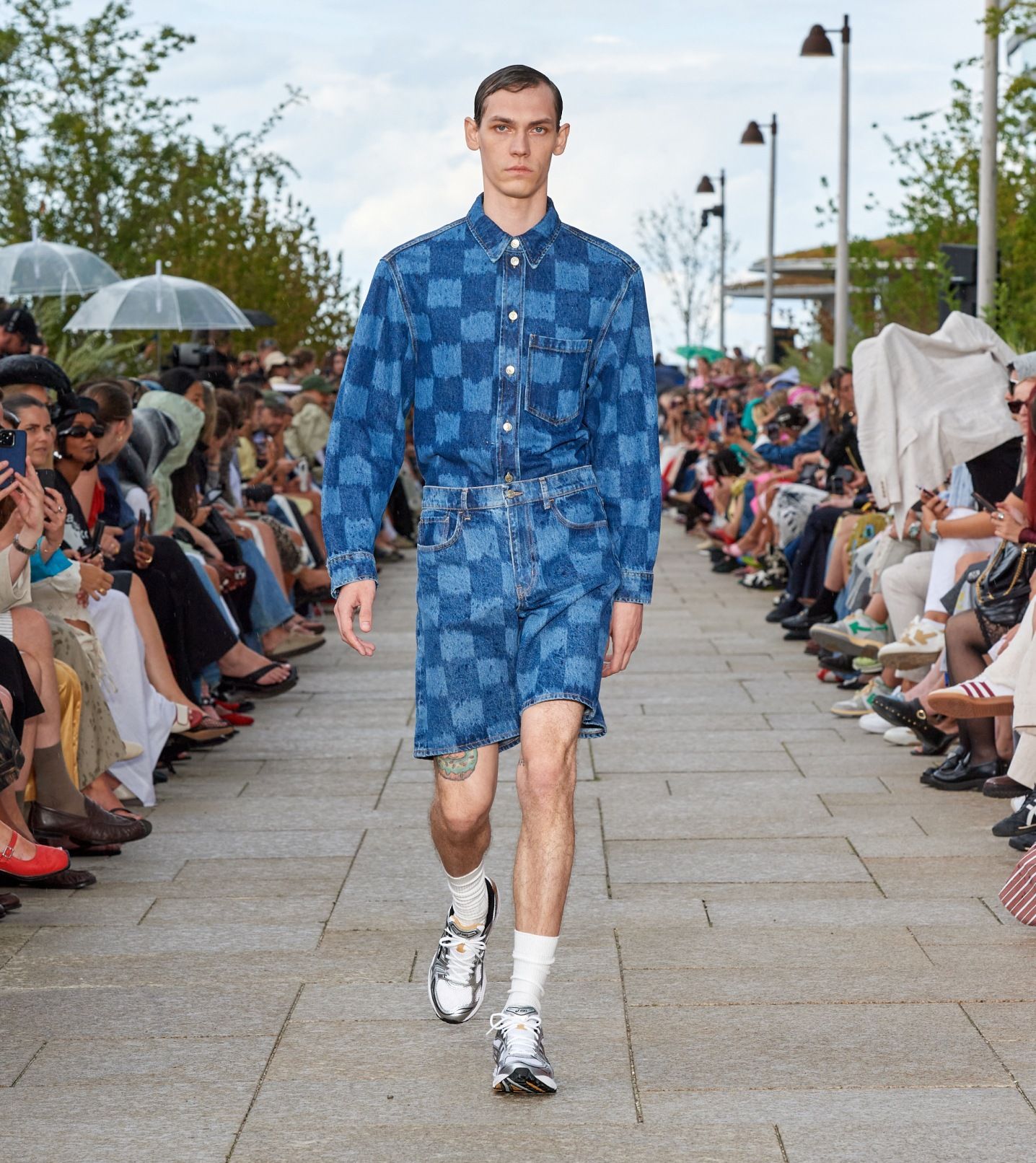 A mModel walking on the catwalk of the fashion show dressed in Jäkälä patterned denim shorts and shirt.