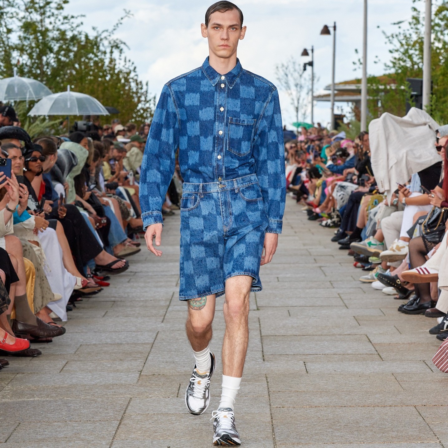 A mModel walking on the catwalk of the fashion show dressed in Jäkälä patterned denim shorts and shirt.
