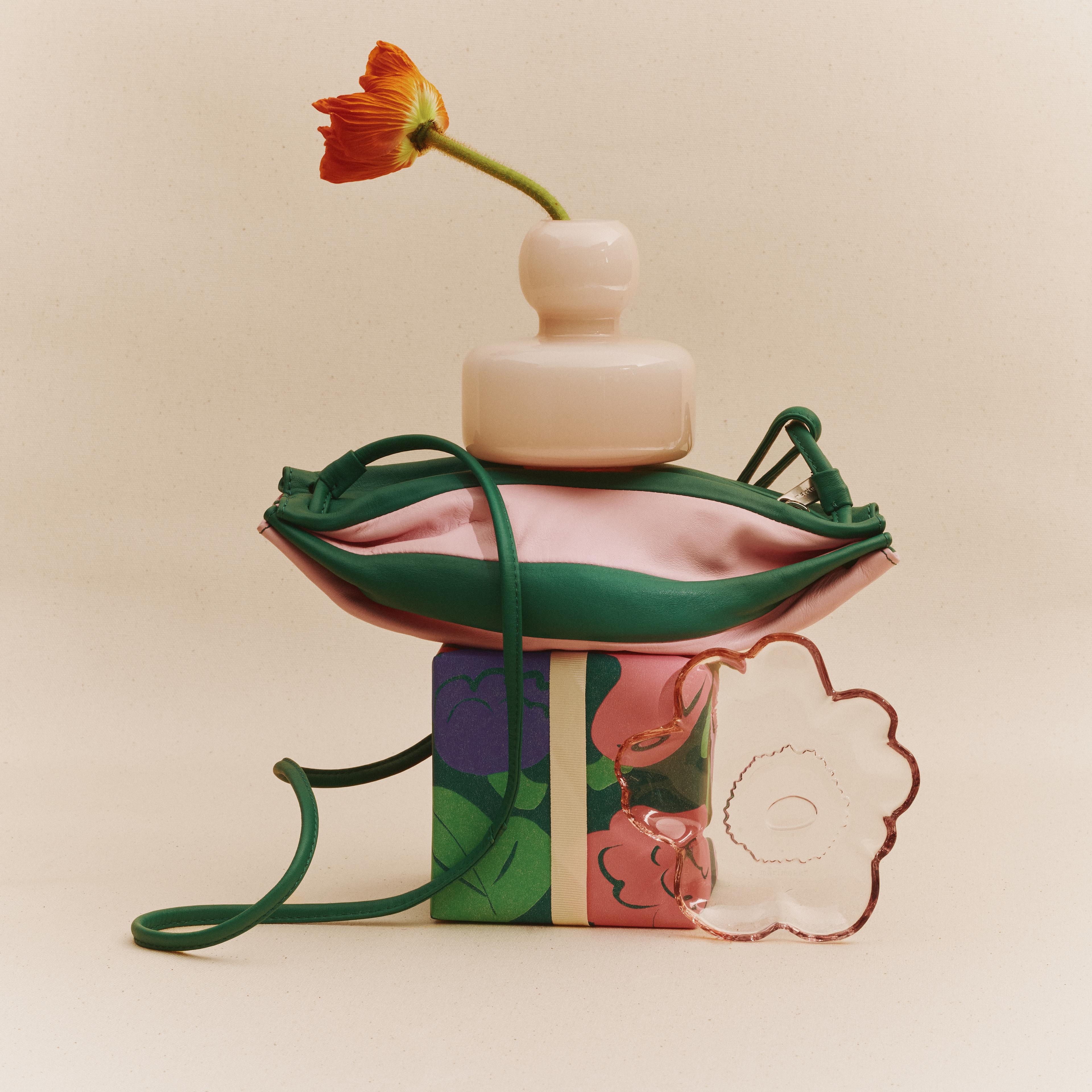 Placed on top of the gift box is a green-pink Karla bag in the shape of a tower, with a Flower vase on top; next to the ensemble is a translucent Unikko plate against a light beige background.