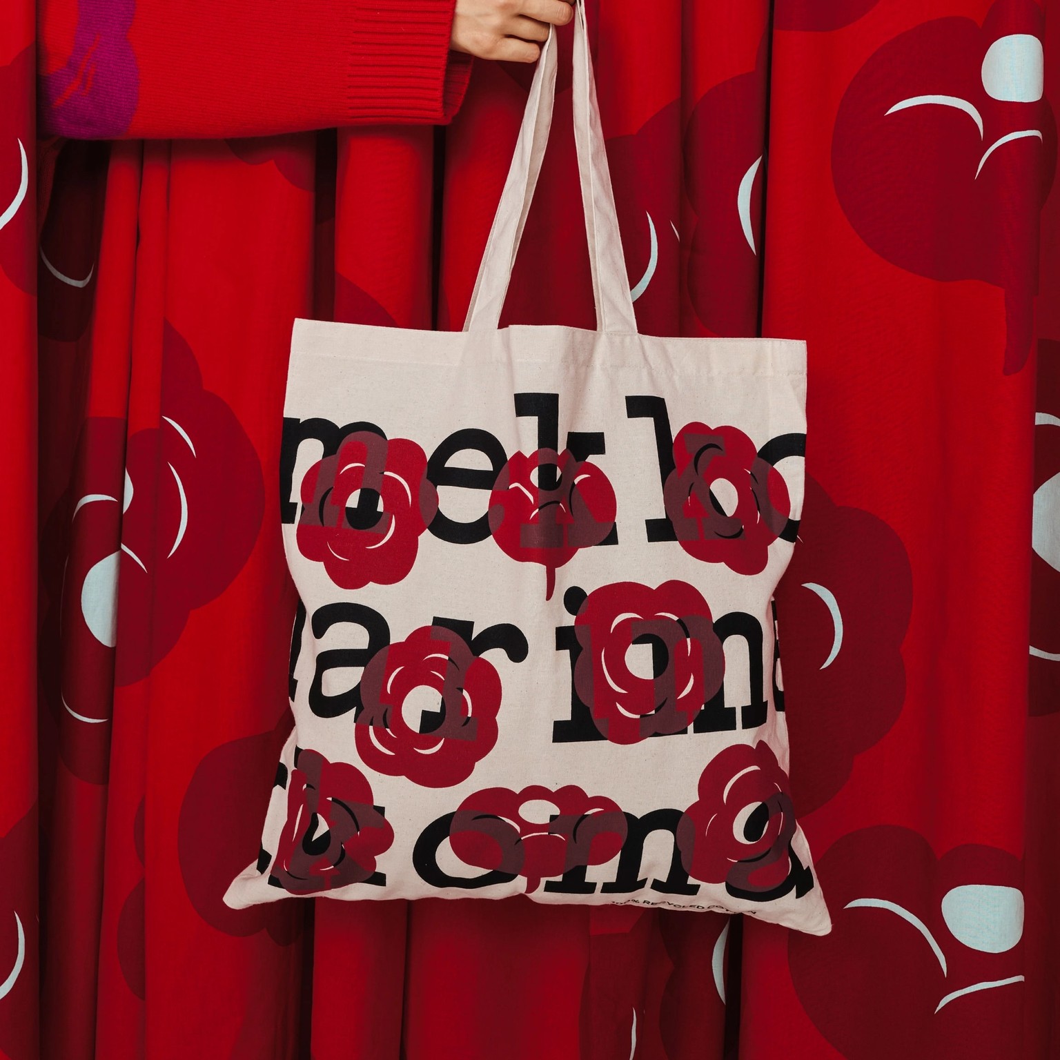 Käsi pitämässä Marimekko-logokassia, jossa punaisia kukkakuviota.