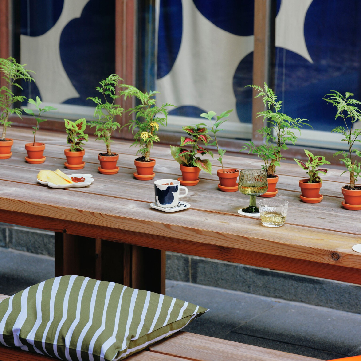 Bar Unikko's terrace is furnished with wooden tables and benches. The tables are decorated with small green plants in pots and wine and cheese in Marimekko items.