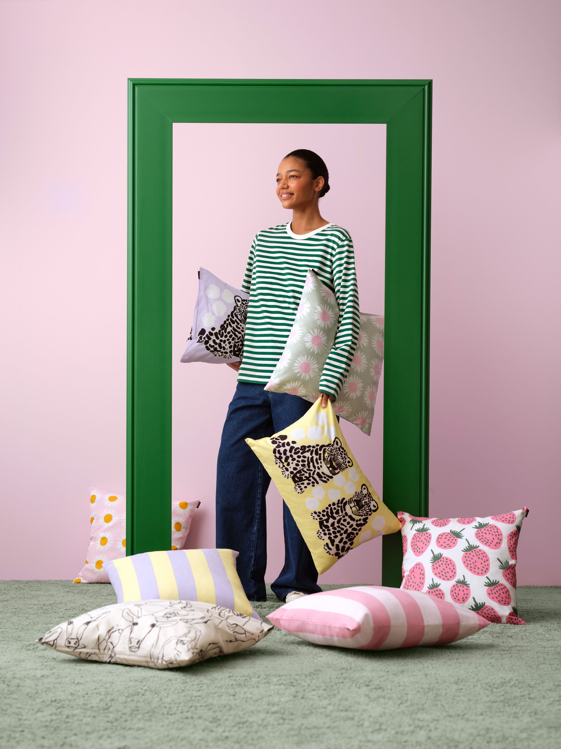 Mallin päällä Tasaraita-pitkähihainen ja hän pitelee sisustustyynyjä eri kuoseissa mm. Korsi, Kaksoset ja Auringonkukka.