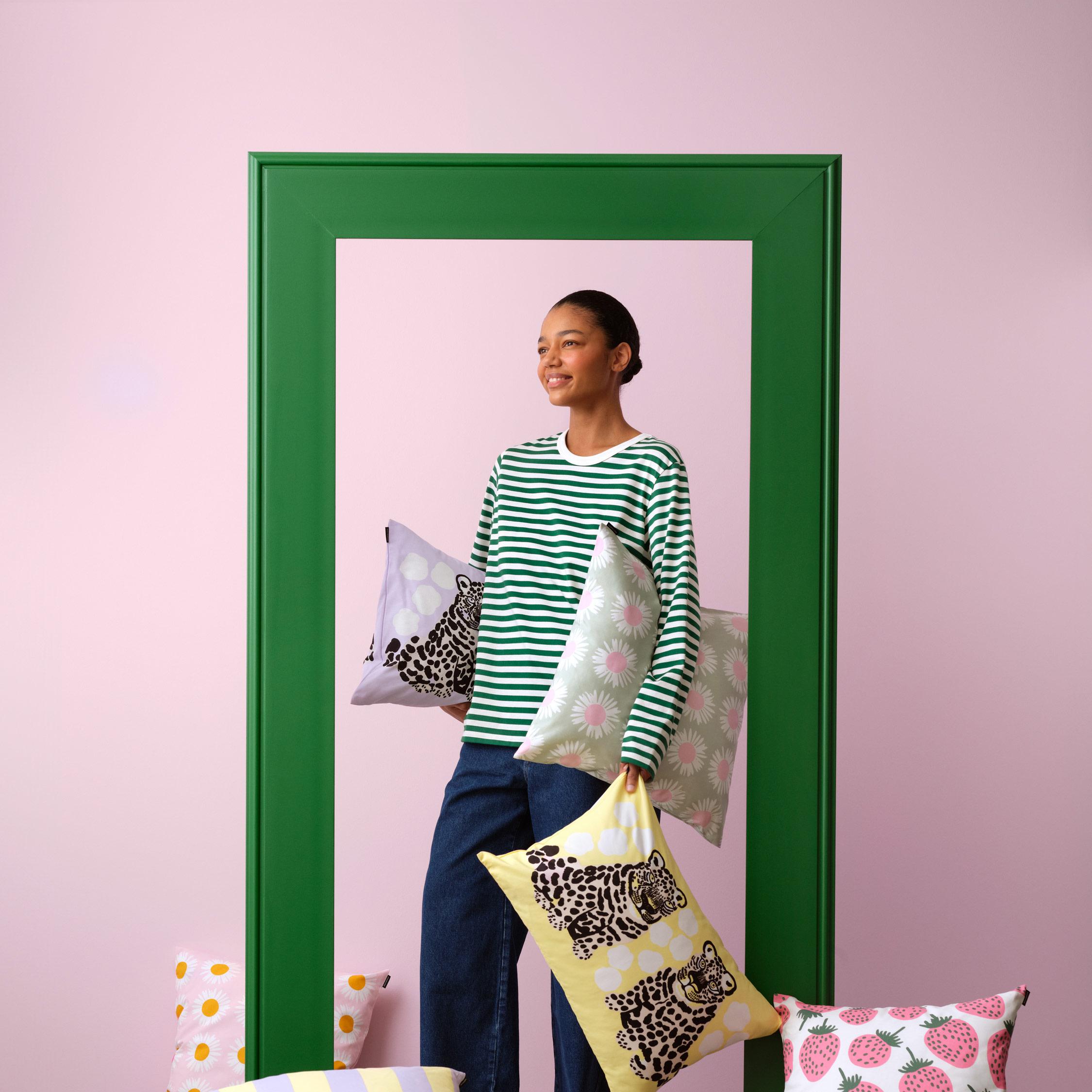 Mallin päällä Tasaraita-pitkähihainen ja hän pitelee sisustustyynyjä eri kuoseissa mm. Korsi, Kaksoset ja Auringonkukka.