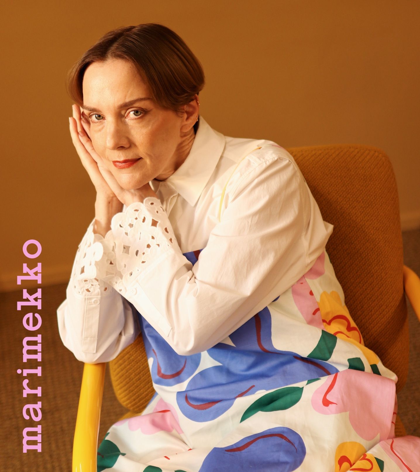 A woman (Maria Veitola) wearing a dress in Kukasta kukkaan print, a shirt with Keidas embroidery and on the image the text ''Marimekko'' vertically in the left bottom corner.