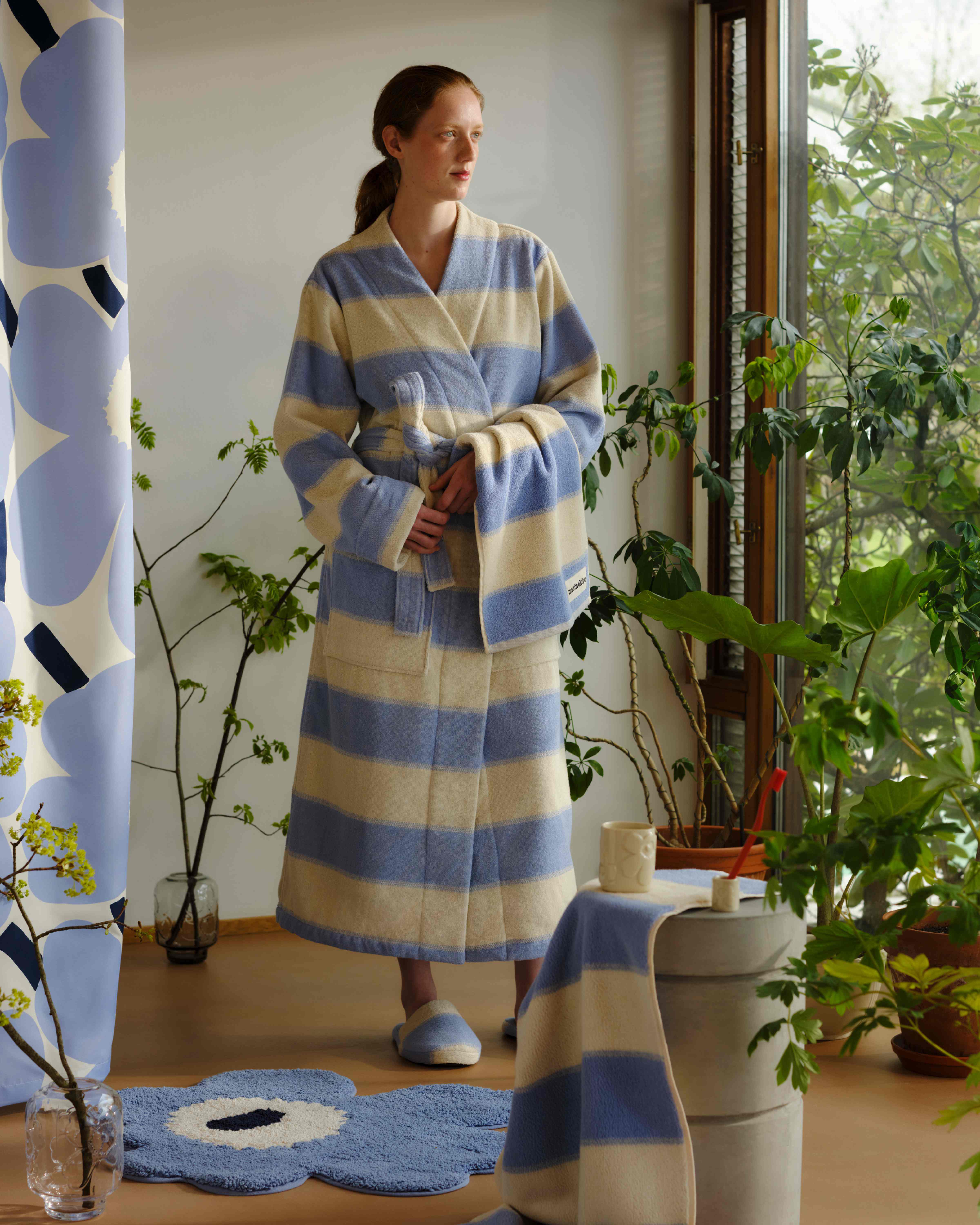 Model in a blue and white striped bathrobe standing in a plant-filled bathroom with a blue Unikko-shaped bath mat and a blue Unikko print shower curtain