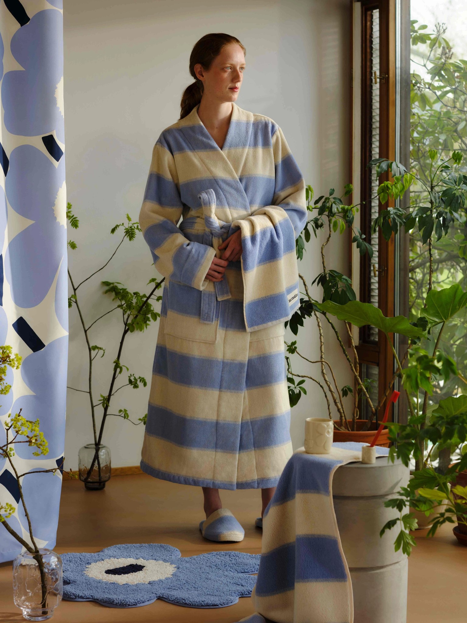 Model in a blue and white striped bathrobe standing in a plant-filled bathroom with a blue Unikko-shaped bath mat and a blue Unikko print shower curtain.