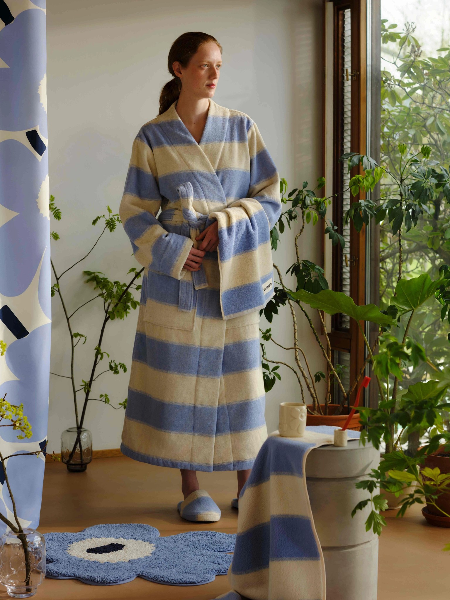 Model in a blue and white striped bathrobe standing in a plant-filled bathroom with a blue Unikko-shaped bath mat and a blue Unikko print shower curtain