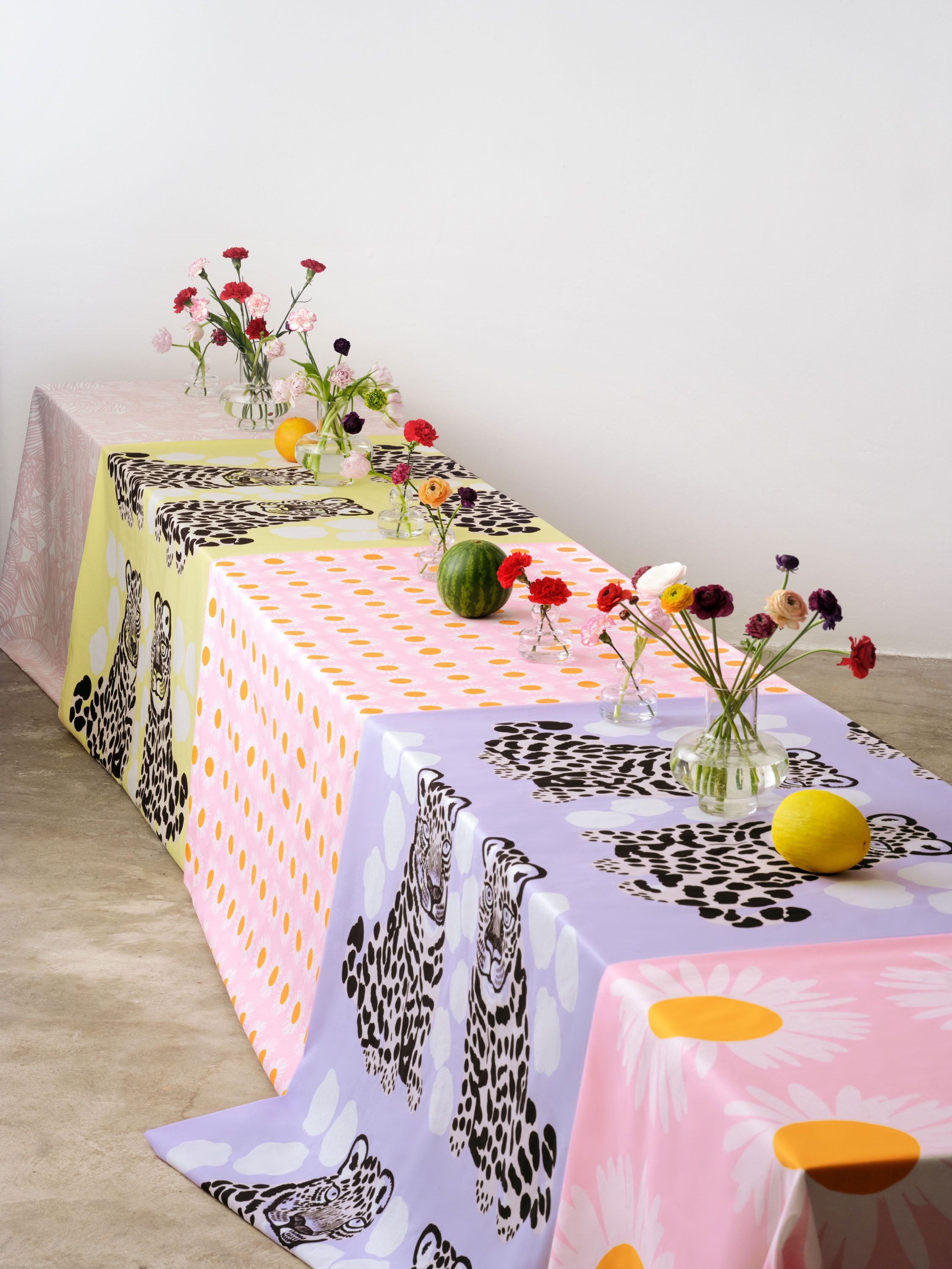 Pitkä pöytä, jossa useita kukkamaljakkoja ja Kaksoset sekä Auringonkukka kankaita pöytäliinoina.