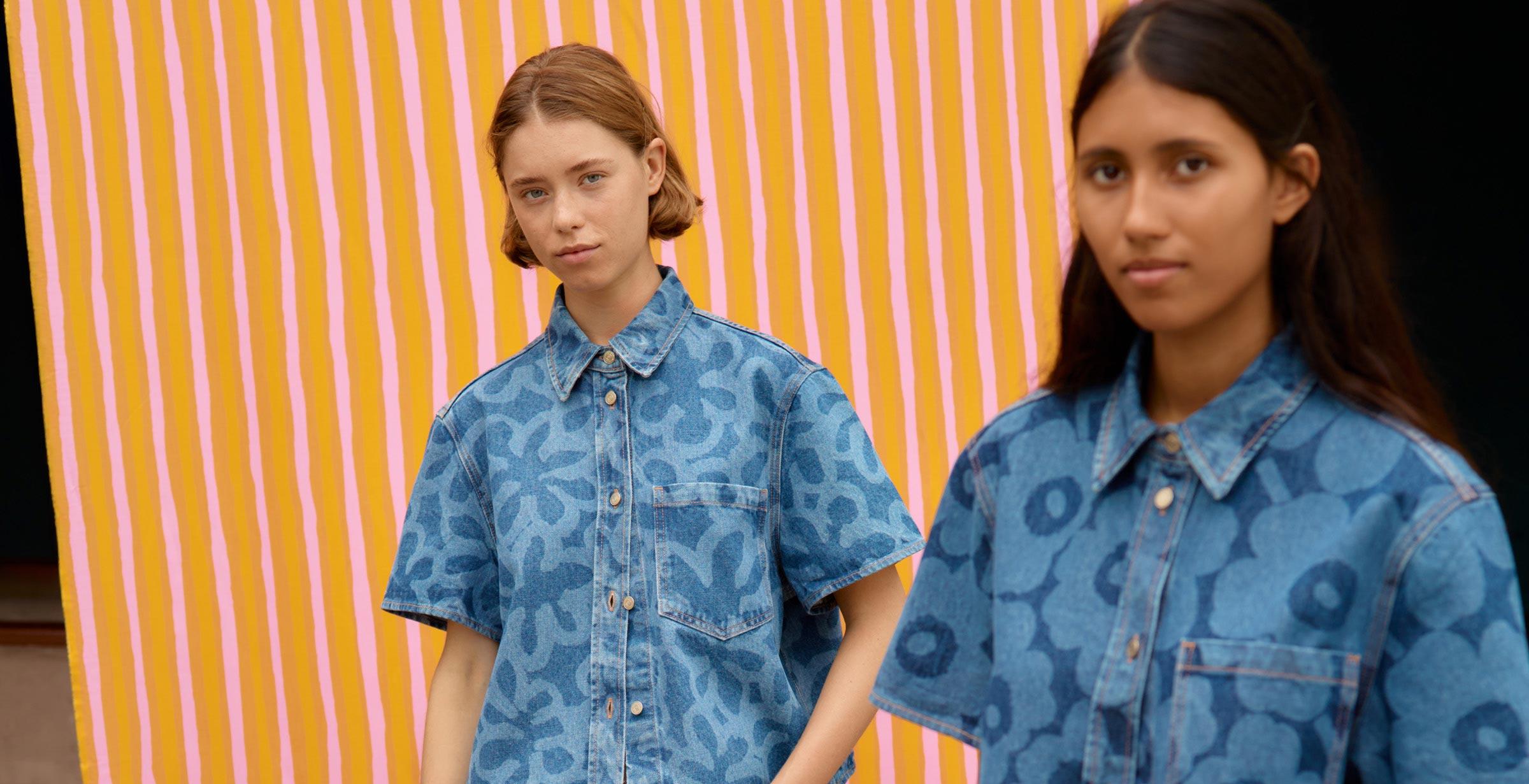 Two models posing in front of orange and pink Piccolo print fabric and wearing a denim tops and skirts in Kukka and Unikko prints.