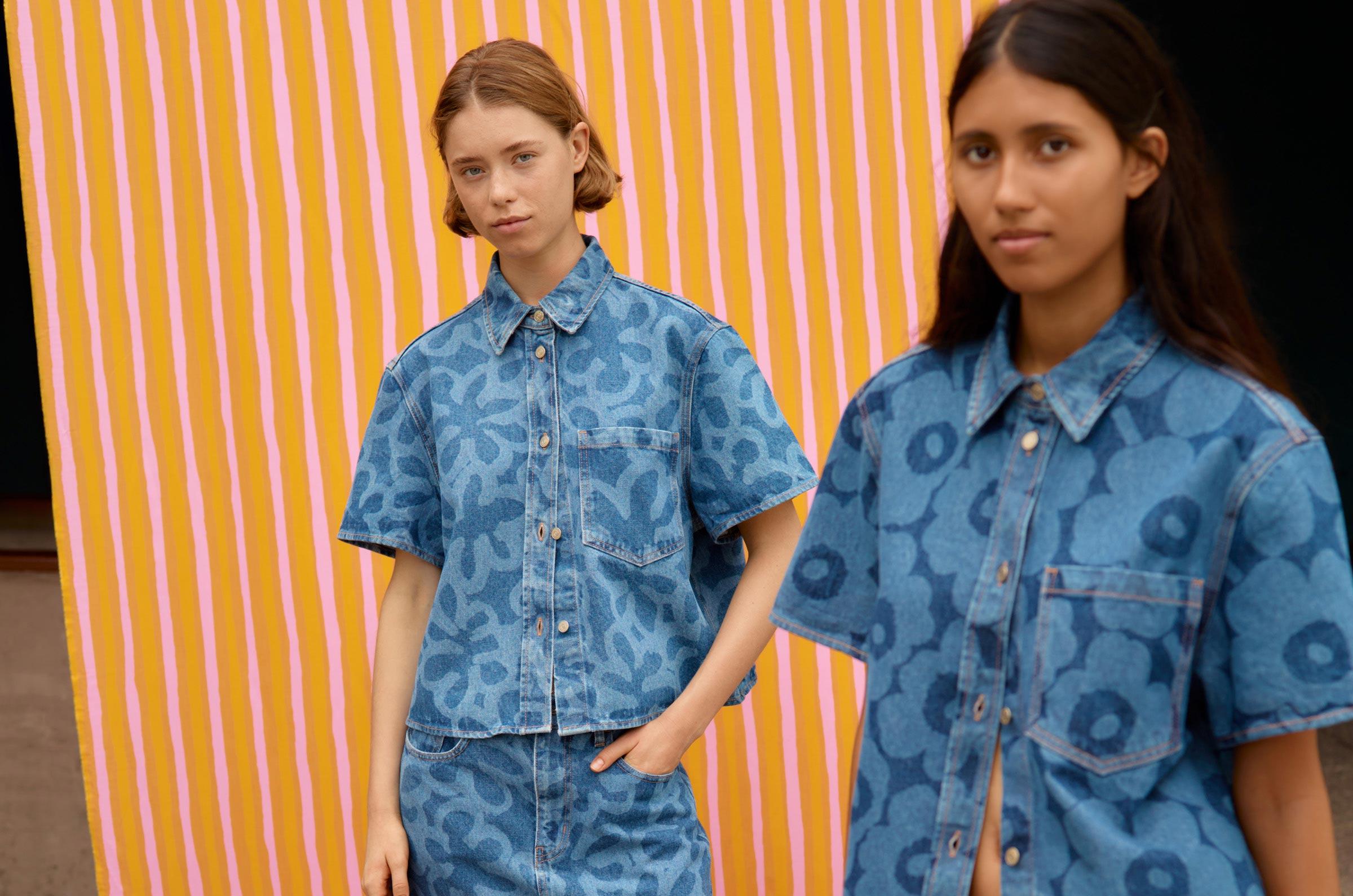Two models posing in front of orange and pink Piccolo print fabric and wearing a denim tops and skirts in Kukka and Unikko prints.