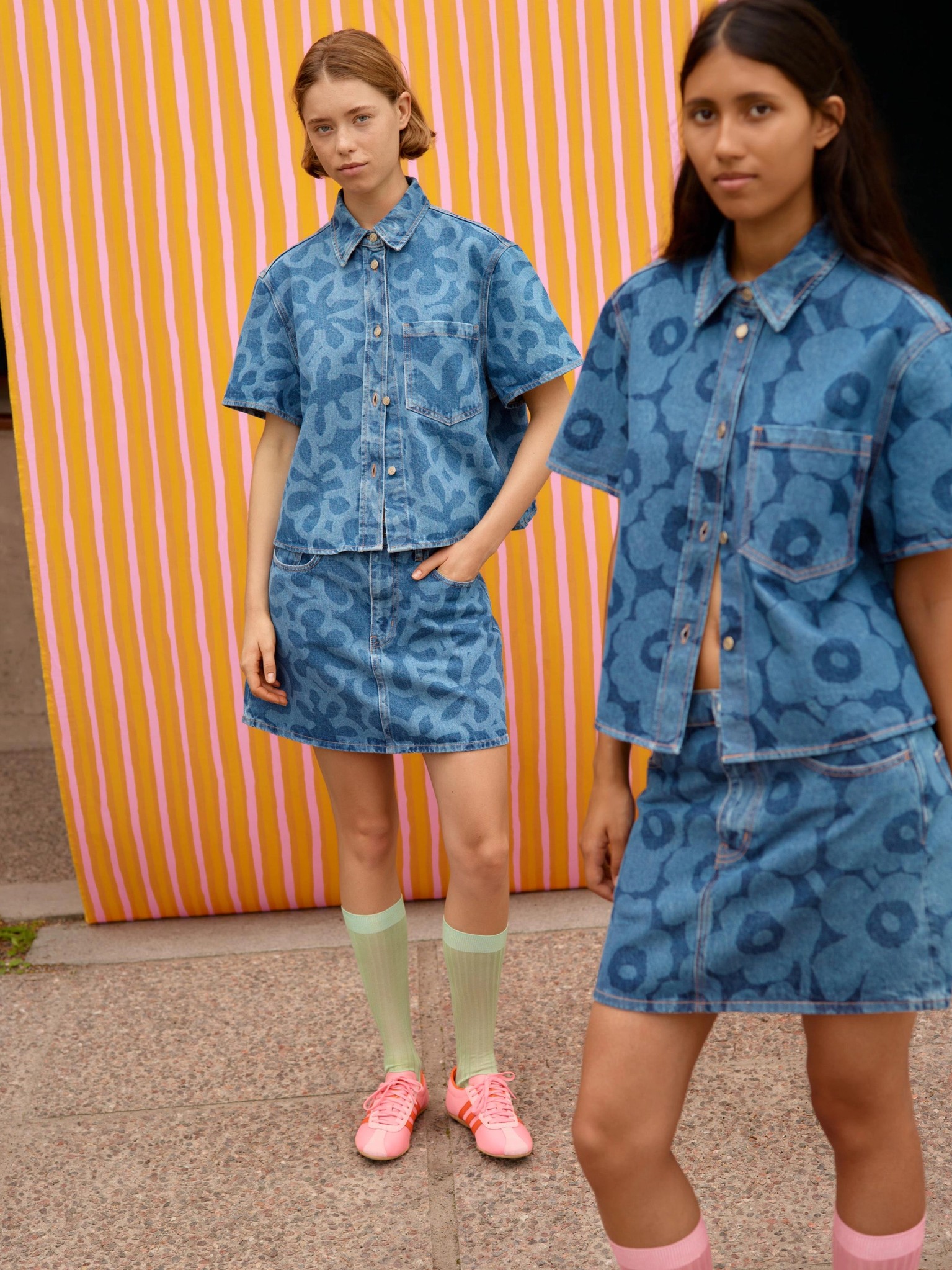 Two models posing in front of orange and pink Piccolo print fabric and wearing a denim tops and skirts in Kukka and Unikko prints.