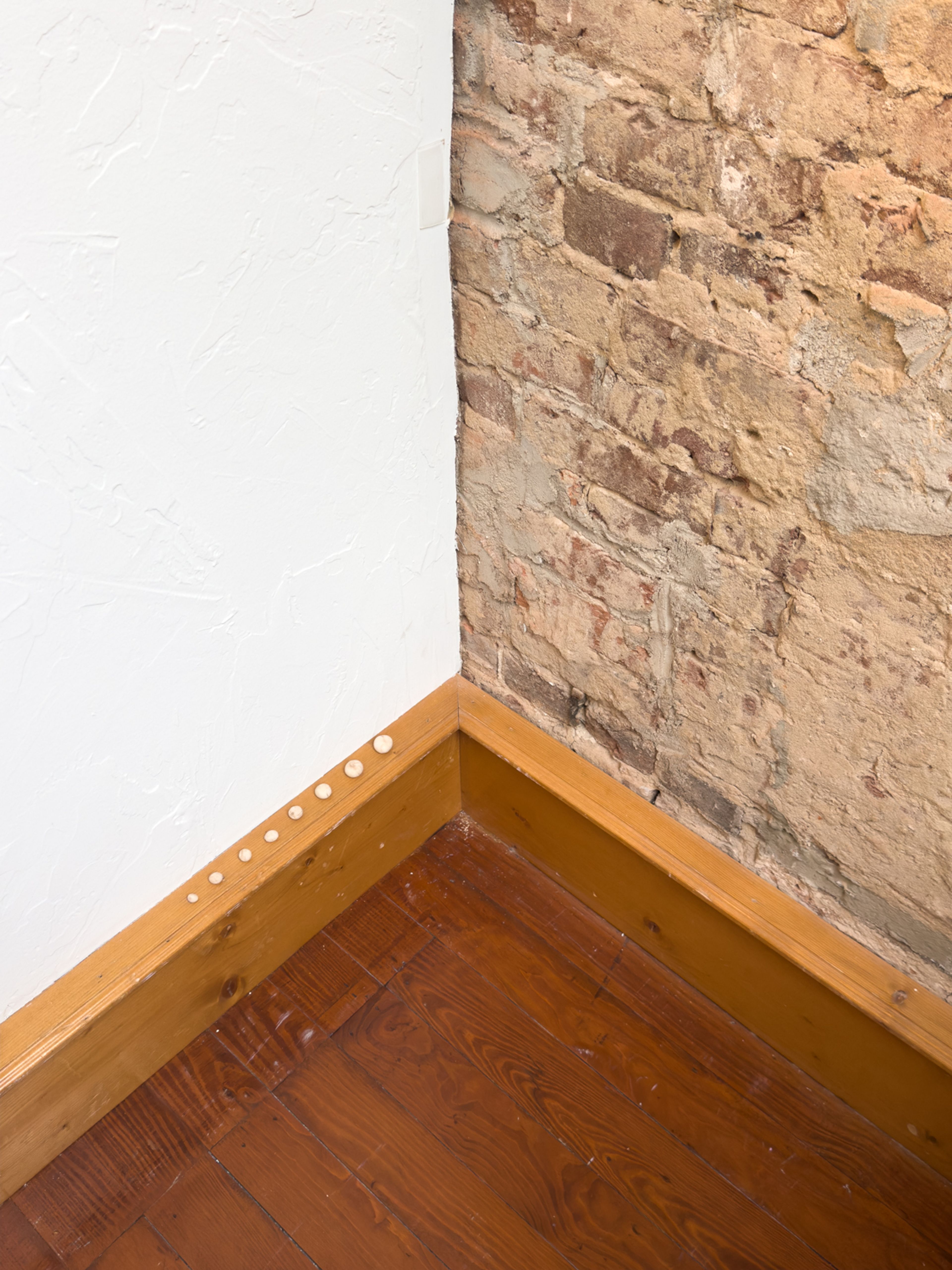 detail photo of the floor at a corner where white wall meets brick wall with wooden trip and dark wood floors. balancing on the wooden trim is a series of small, white, clay balls in ascending order according to size