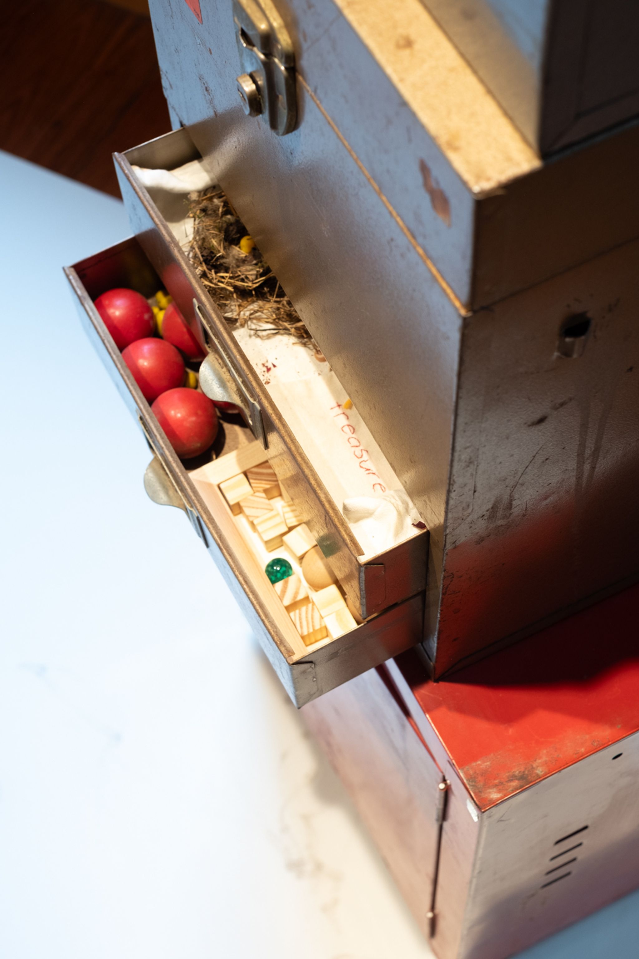 detail of a metal filing cabinet with two drawers open that include various small red orbs, wooden blocks, marbles, and a nest