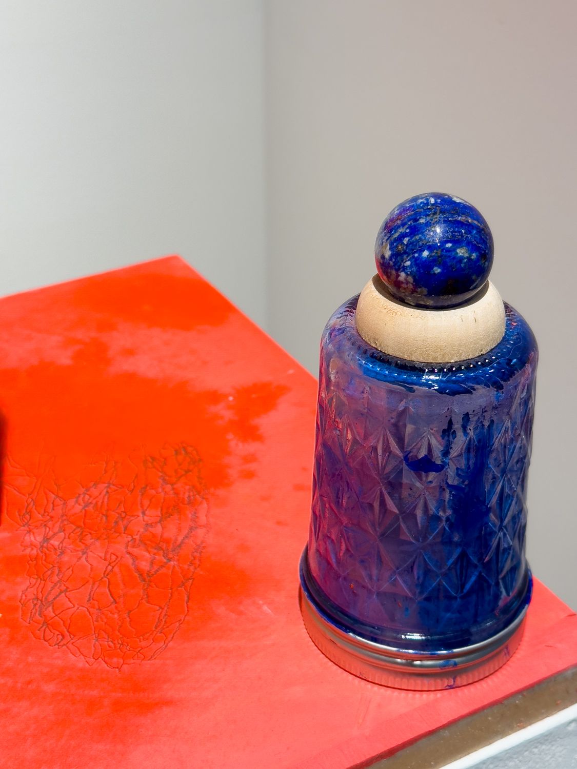 A textured blue glass jar filled with blue pigment is capped by a light wood sphere and a lapis lazuli sphere, resting on a textured red surface.