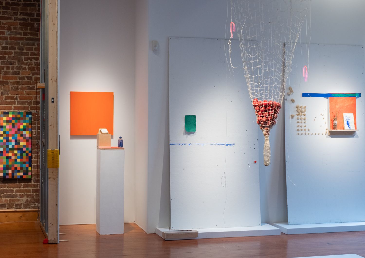 a gallery showing part of a gridded colorful large painting on a brick wall, orange square painting, objects on a pedastal, two white panels with small colorful pieces leaned against a wall, a handmade net with red orbs