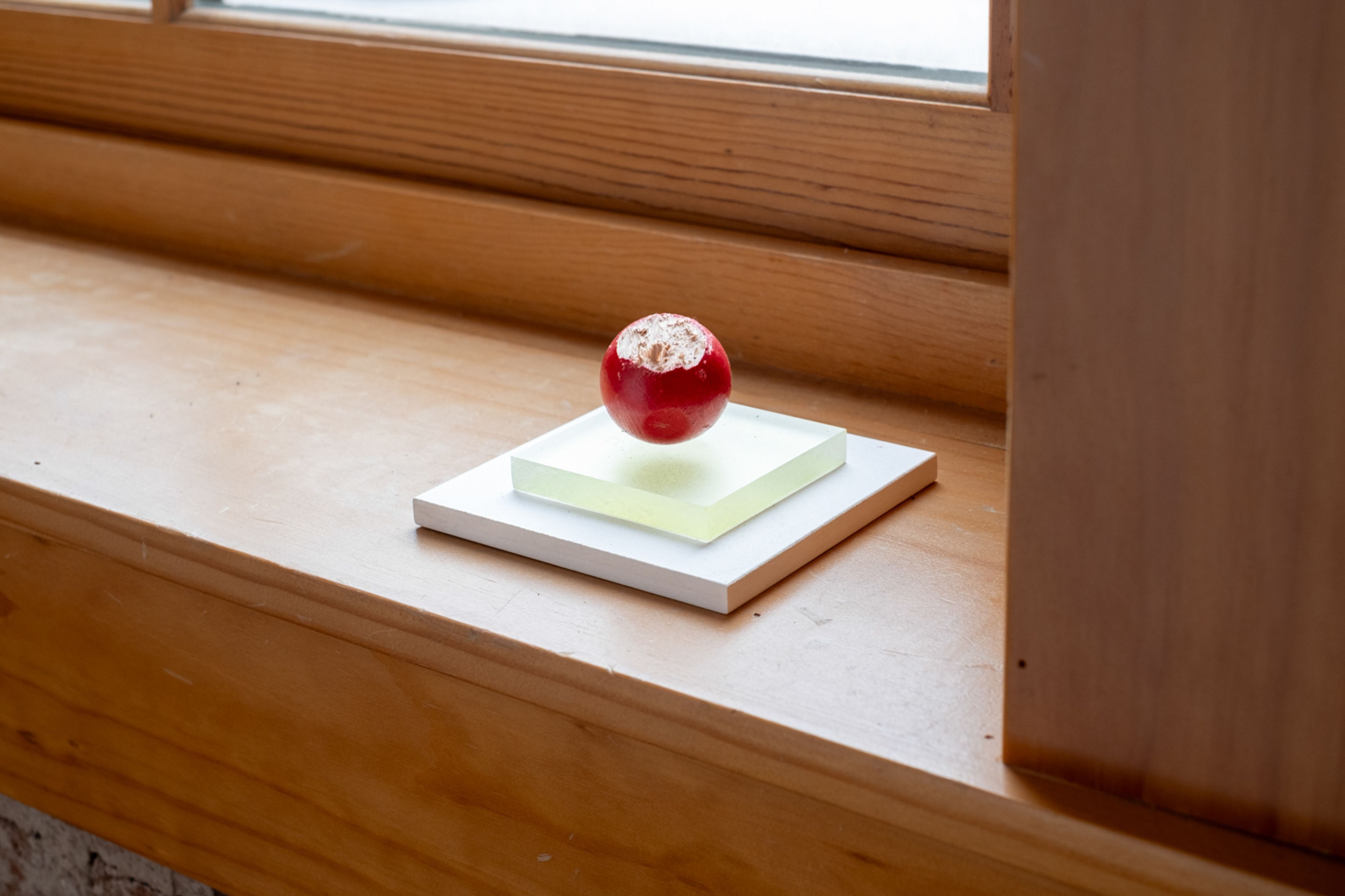 a red wooden orb with a bite out of the top sits on two stacked square supports the bottom is white and top is a clear green. all of this sits on the windowsill in the gallery
