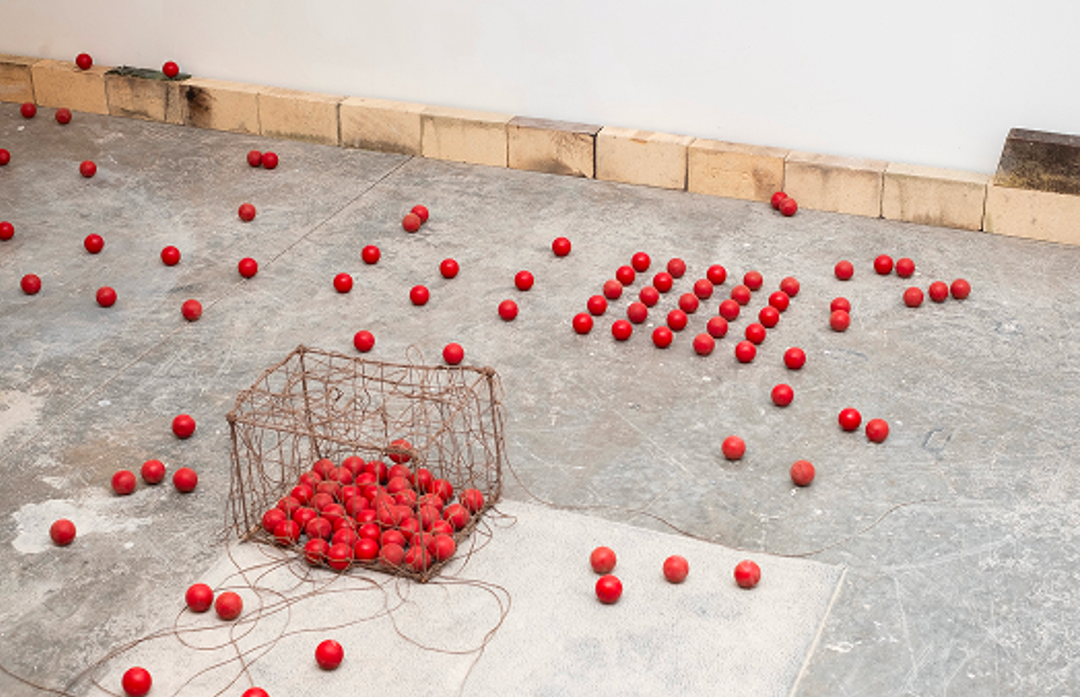 An art installation featuring numerous red spheres on a concrete floor, some in a wire basket, others arranged in neat rows.