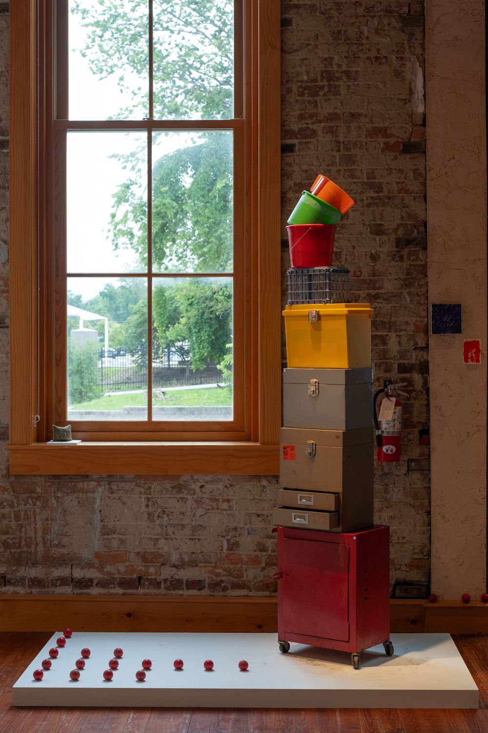 a sculpture on a white plinth next to a tall rectangular window in the gallery. on the plinth on the left is a collection of small red orbs, on the right is a tall, column like sculpture made of stacked metal and plastic bins of varying colors