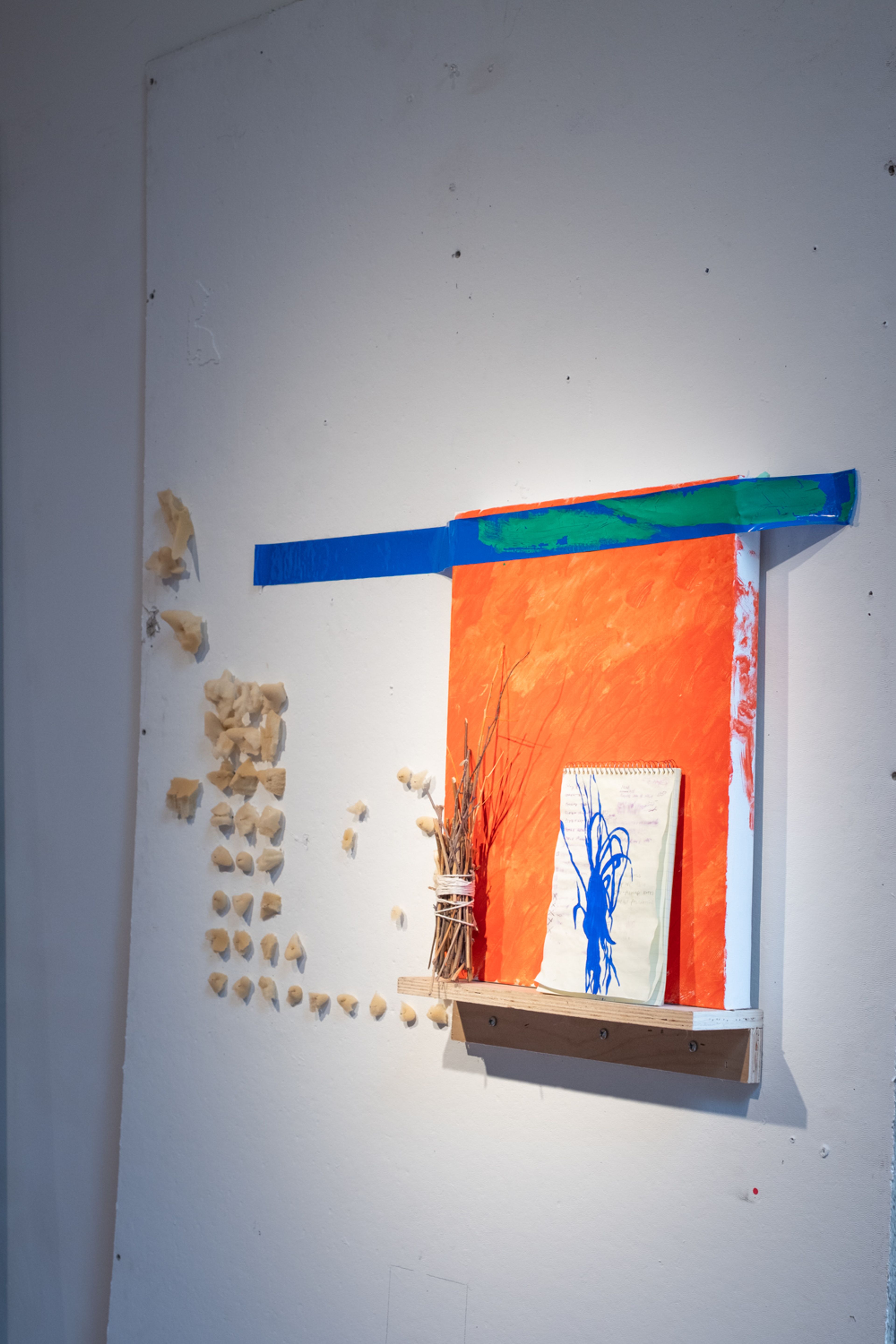 an assemblage on a white panel including tan bits of foam pinned to a wall in a grid pattern, a wood shelf holding an orange painting, supported by blue tape with green paint on it, in front of the painting is a bundle of sticks and a small green notebook with a blue silhouetted painting of the bundle of sticks overlaying a smudged to-do list