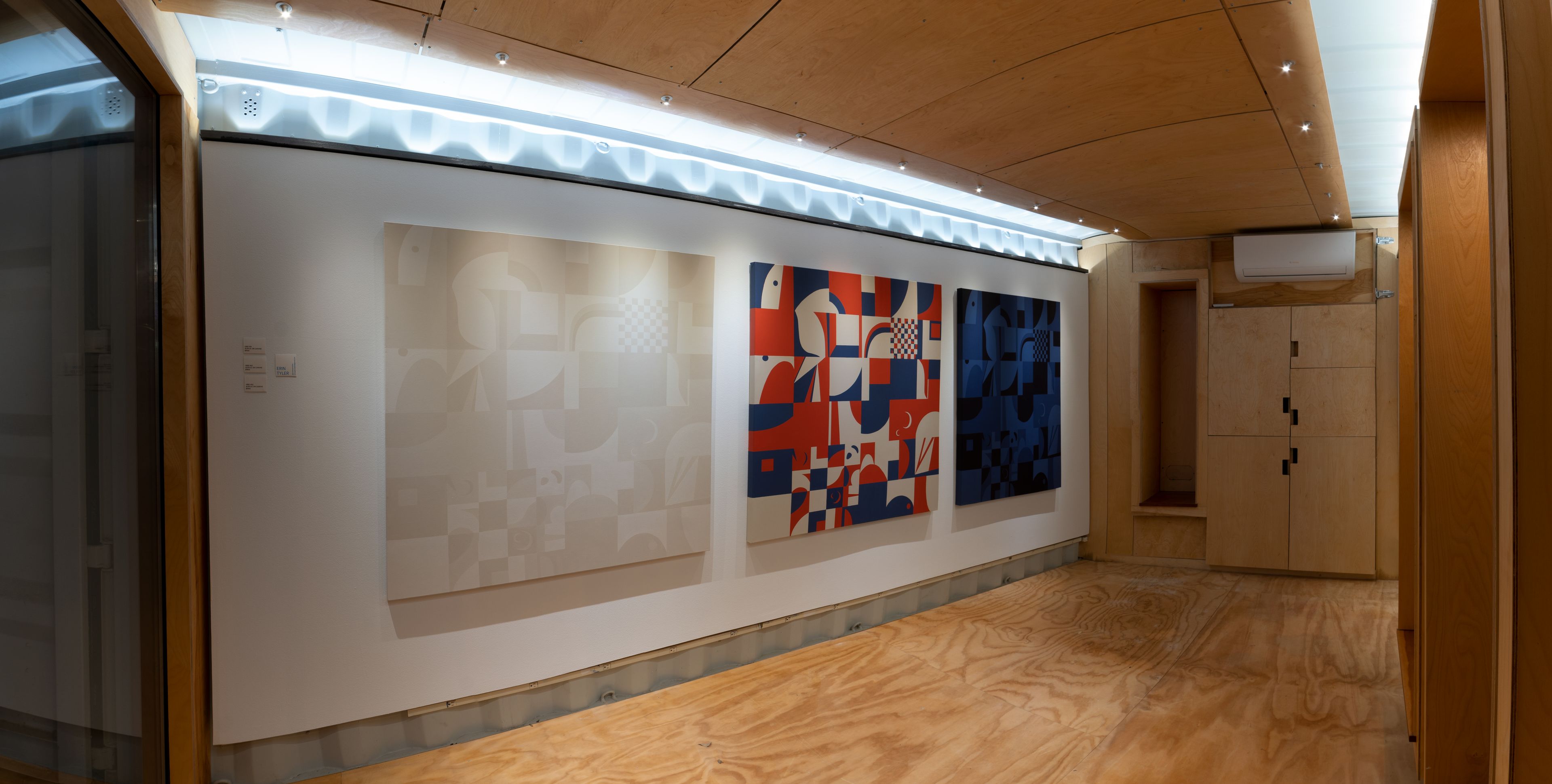 a small shipping container gallery with wood floors and white walls hold three geometric grid large paintings side by side