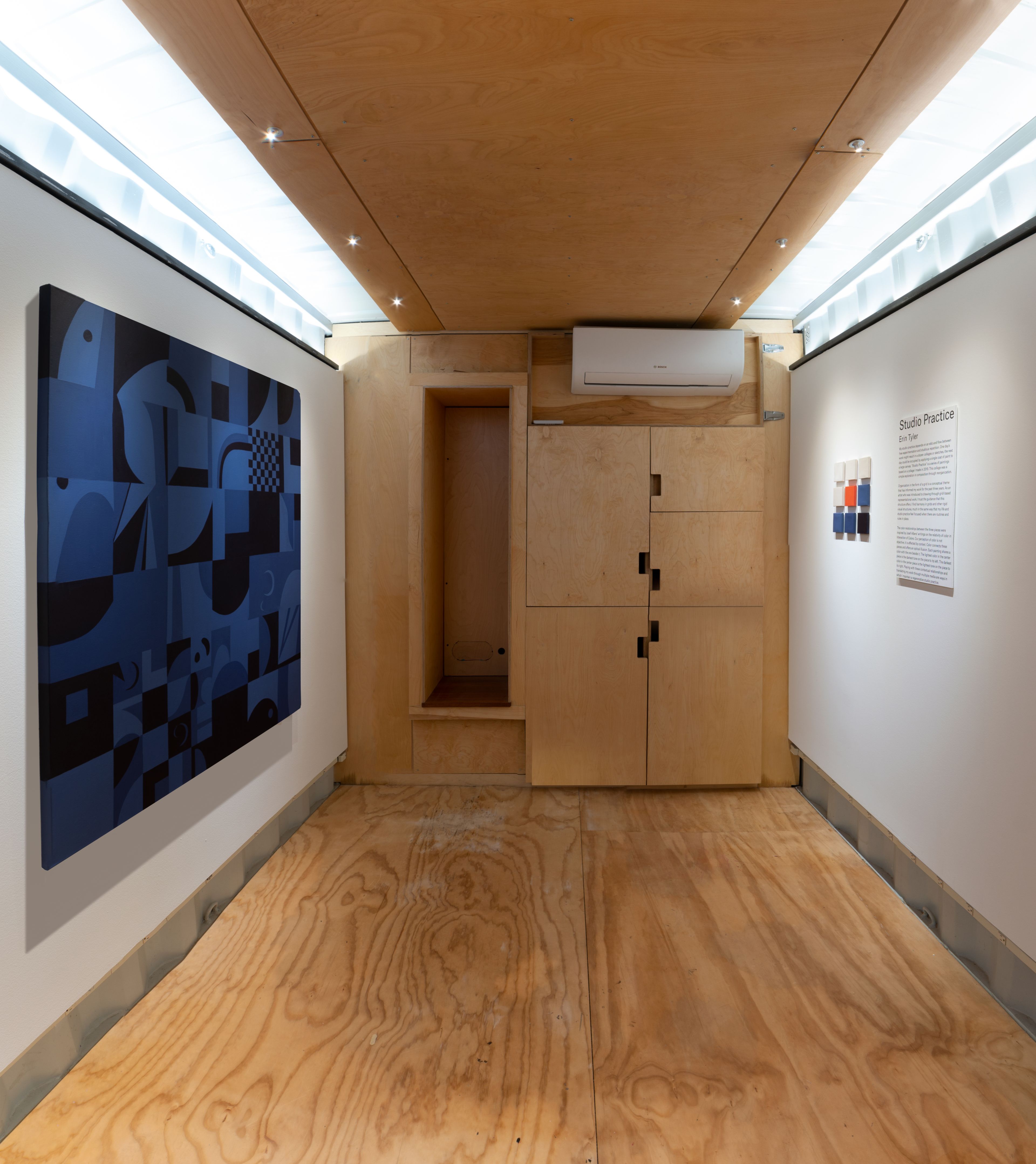 a small shipping container gallery white wood floors and white walls and wood cabinets with a large geometic gridded blue painting and small red, blue and white panels next to a printed artist statement on the right