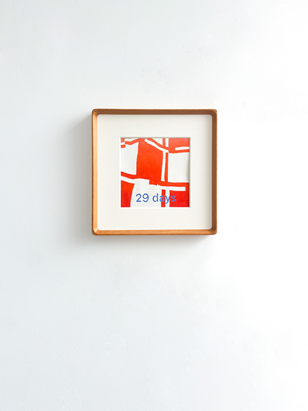A square artwork in a light wooden frame with a white mat, featuring red abstract shapes and the blue text "29 days", hanging on a white wall.
