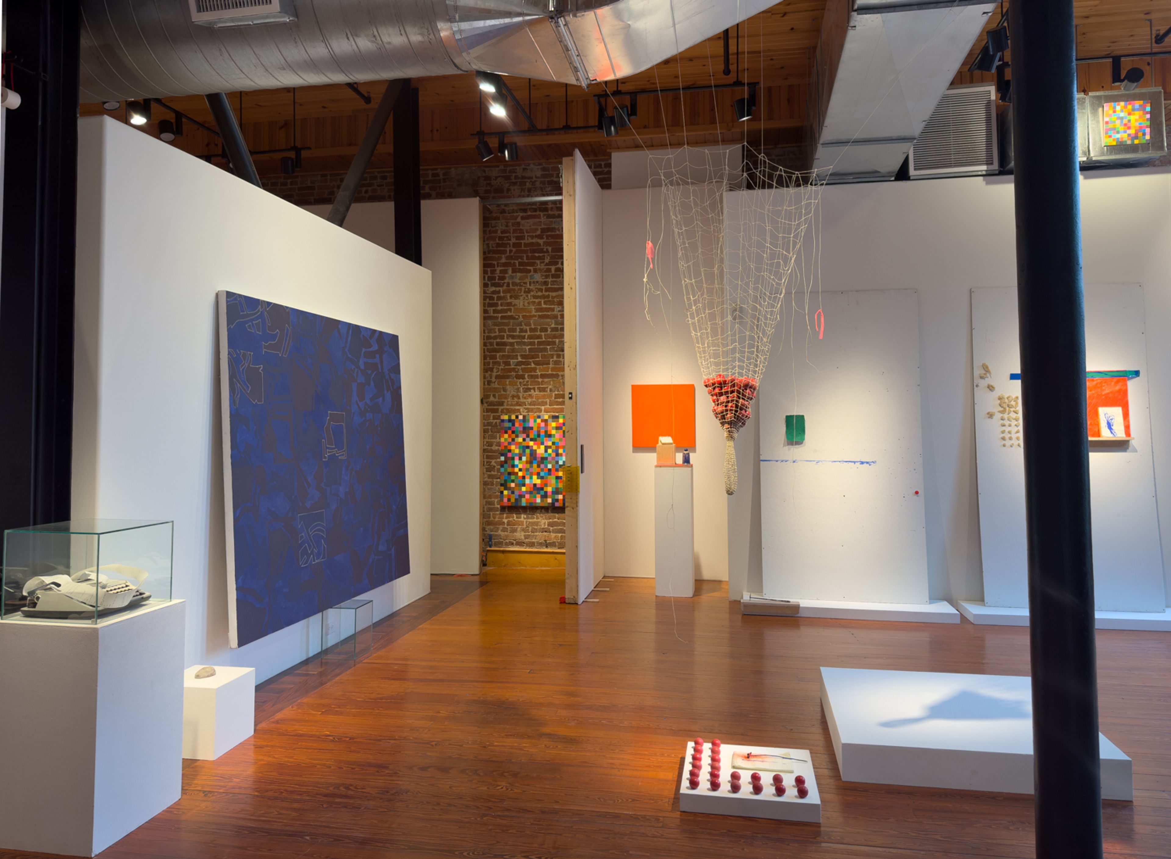 An art gallery with wooden floors and white walls displaying contemporary artworks, including a large blue painting and a hanging net sculpture.