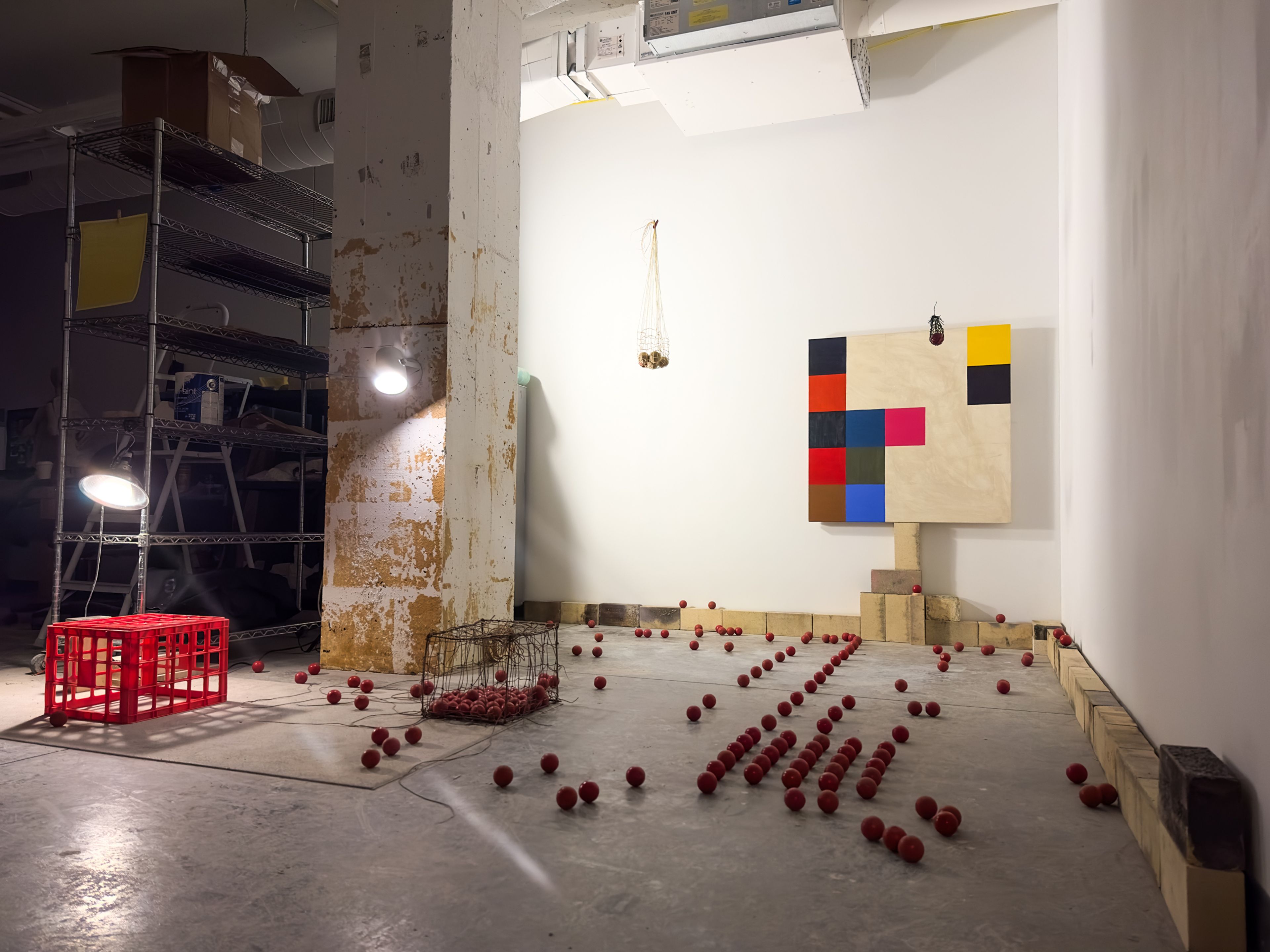 An installation view in a studio or gallery space shows a geometric painting mounted on the wall, composed of colored squares on a tan-toned surface. Below it, a line of wooden blocks runs along the wall. Dozens of small red spheres are scattered and arranged in loose lines across the concrete floor, some contained in a handmade cage. To the left, a red crate and metal shelving with tools and materials sit beside a worn structural column under exposed lighting.