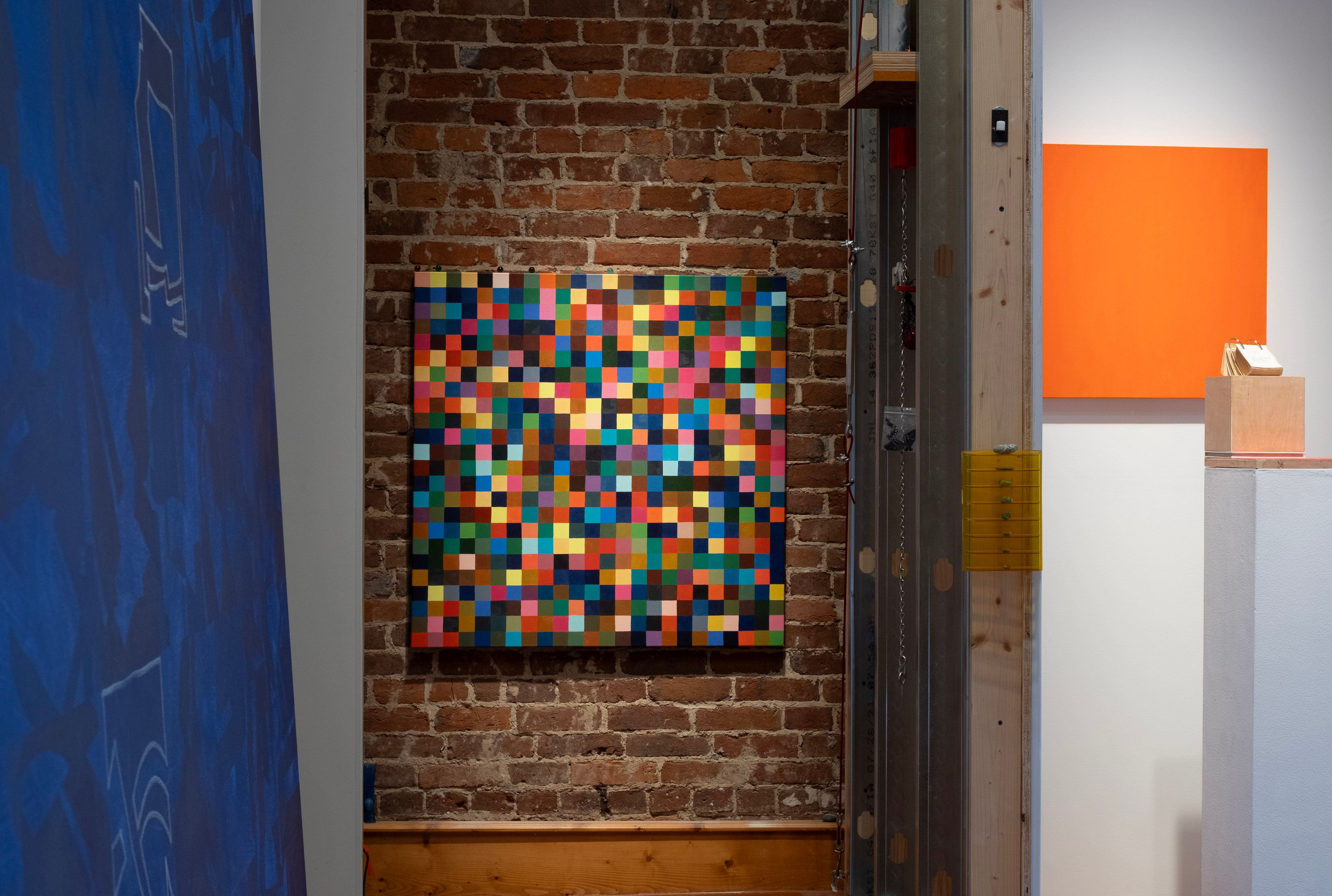 An art gallery features a colorful pixelated painting on a brick wall, an orange square artwork, and a partially visible blue painting.