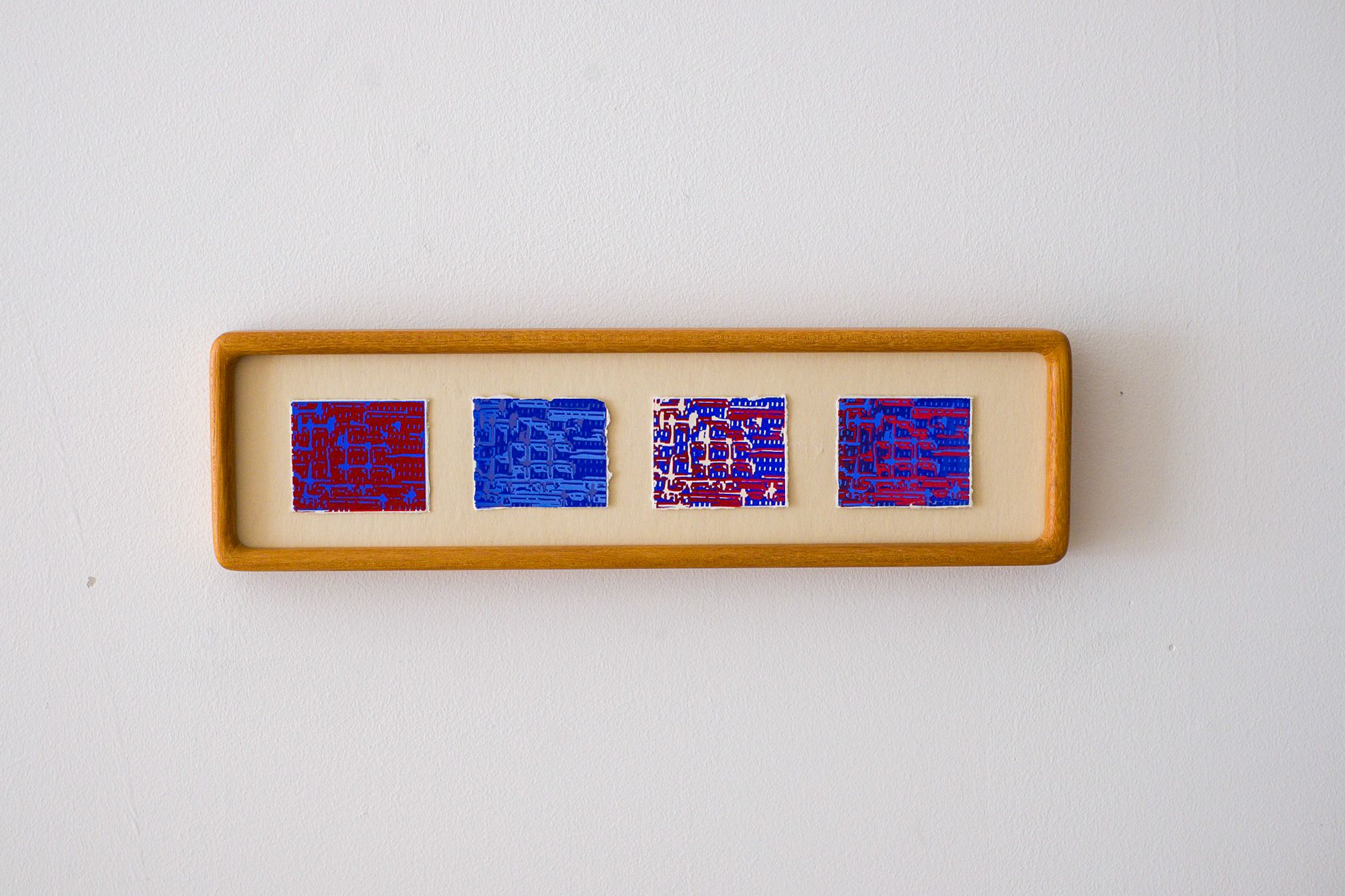 A horizontal wooden frame on a white wall holds four abstract artworks with red, blue, and white patterns.