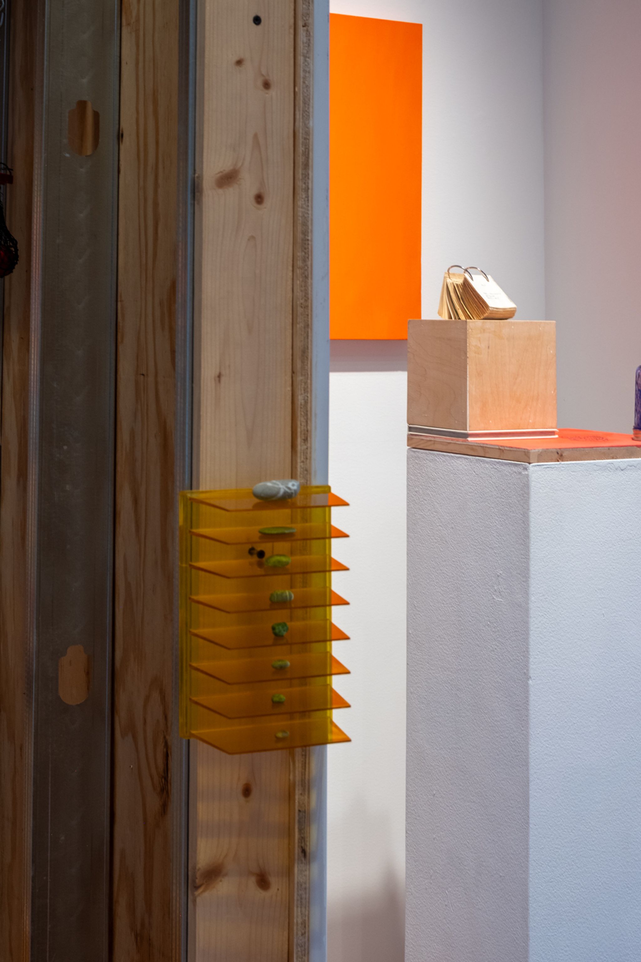 A gallery view showing a wooden wall with an orange tiered shelf holding small green items, an orange rectangular artwork on a white wall, and a wooden cube displaying fanned golden tags on a white pedestal.