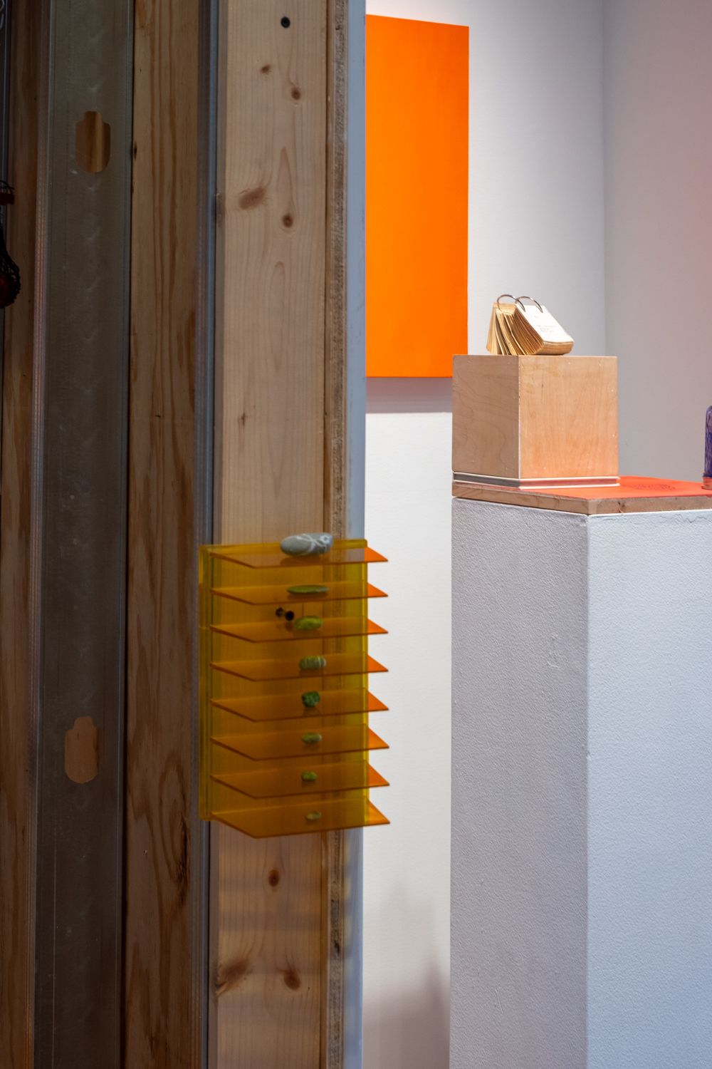 A gallery view showing a wooden wall with an orange tiered shelf holding small green items, an orange rectangular artwork on a white wall, and a wooden cube displaying fanned golden tags on a white pedestal.