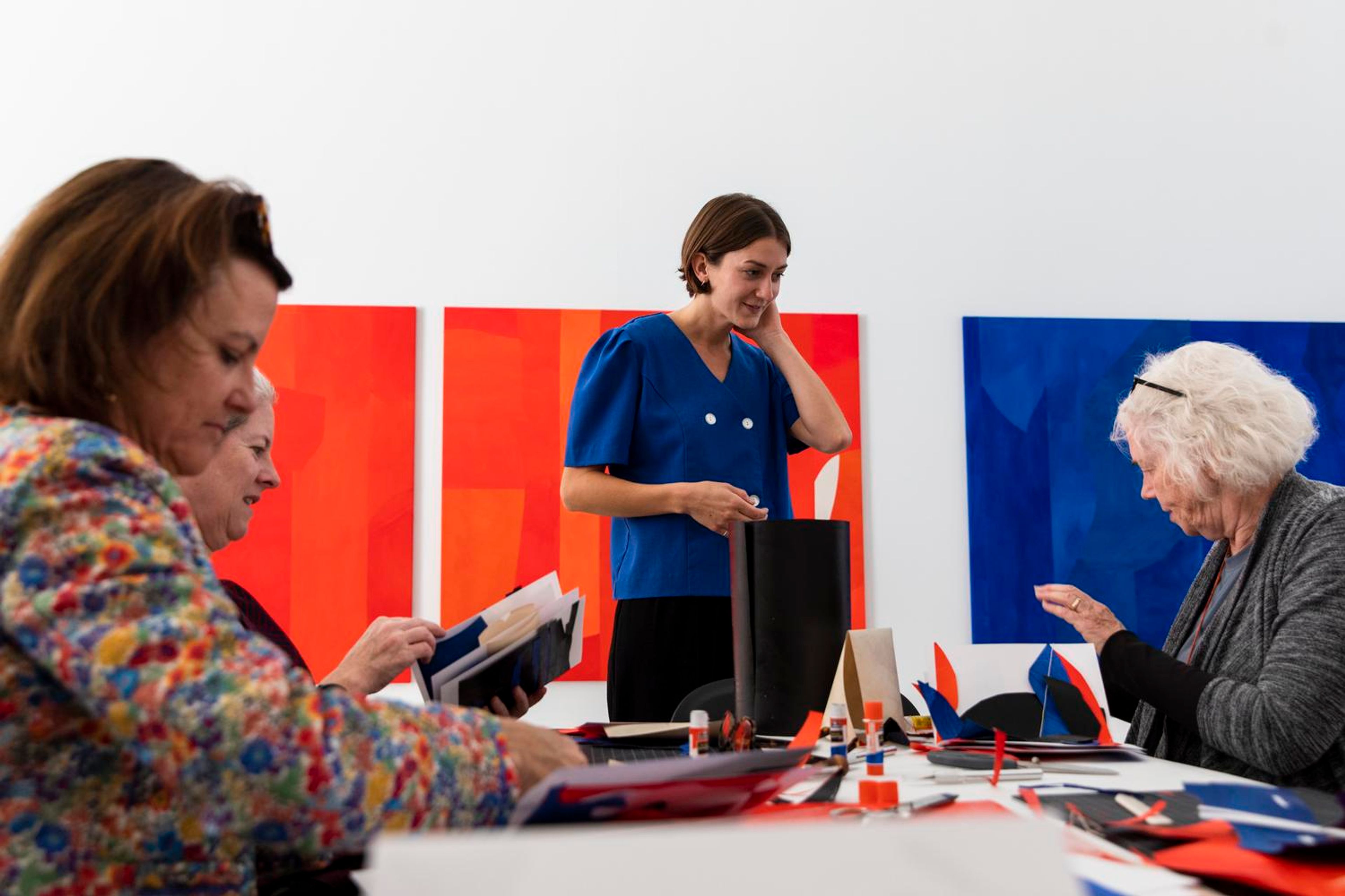 A group of women creating colorful paper collages at a table, with abstract paintings on the wall in the background.