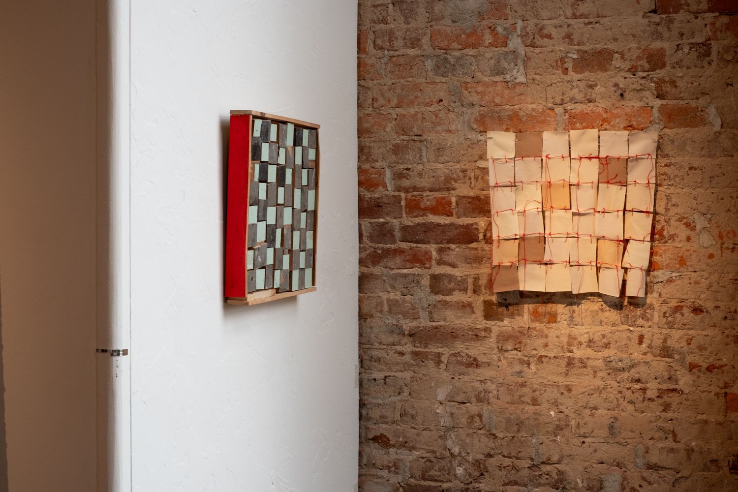 two 12 inch square pieces hang in a corner of the gallery the left most on a white wall showing a red edge and a grey and blue irregular grid made of assembled wood pieces, the right on a brick wall shows small neutral colored squares of fabric stitched together with red string