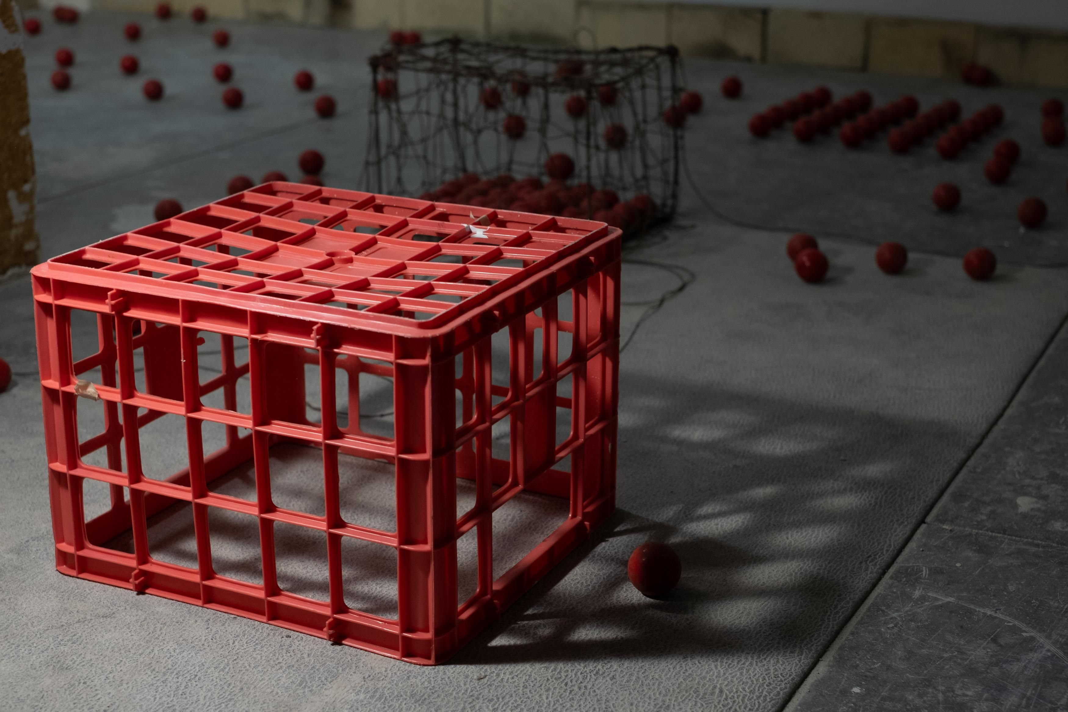 A red plastic crate, numerous red balls scattered on a grey floor, and a wire cage filled with balls in the background.
