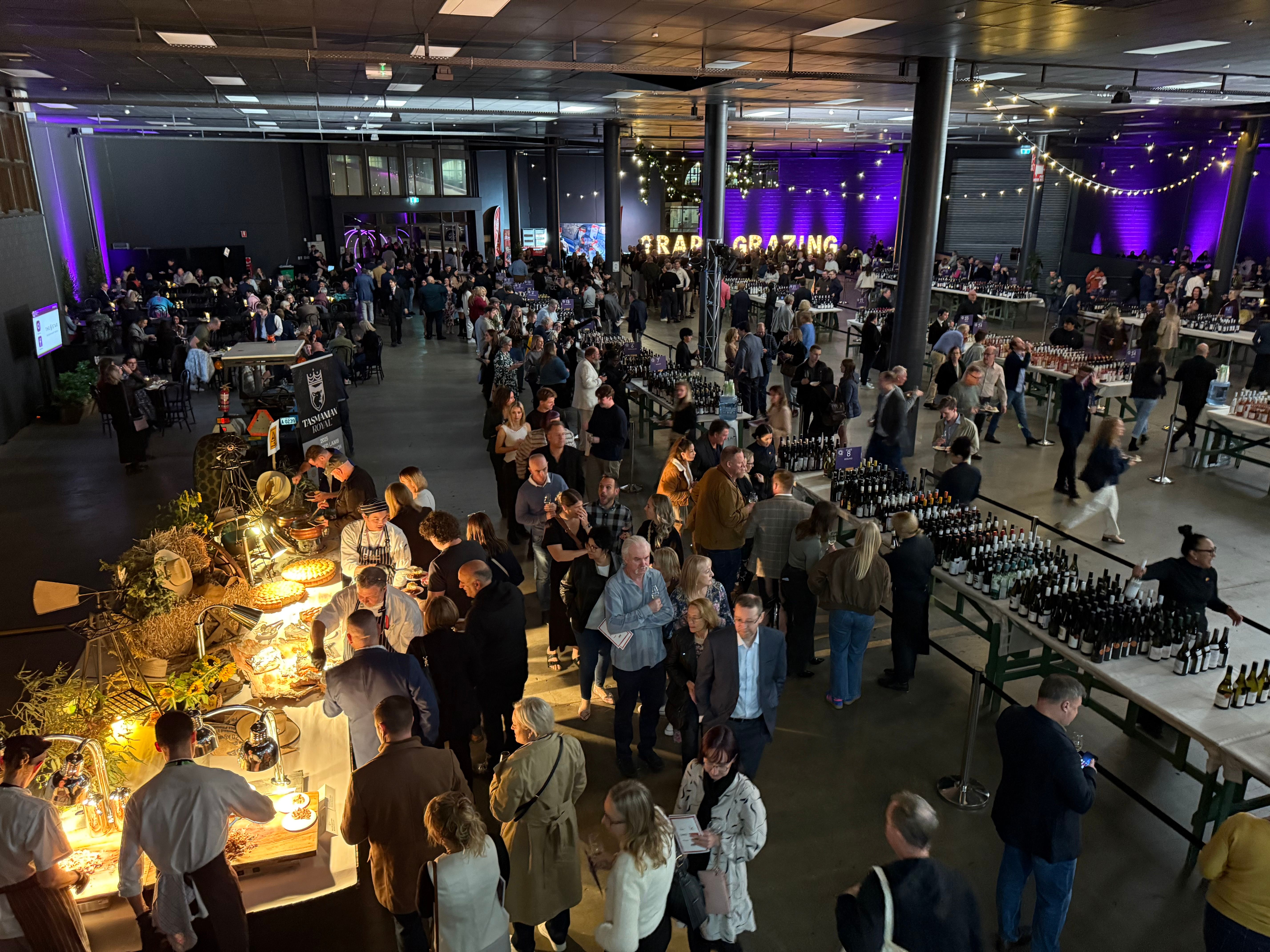 A busy indoor wine tasting event with guests exploring rows of wine bottles, food stations and vendor tables, set beneath warm string lights and purple event lighting.