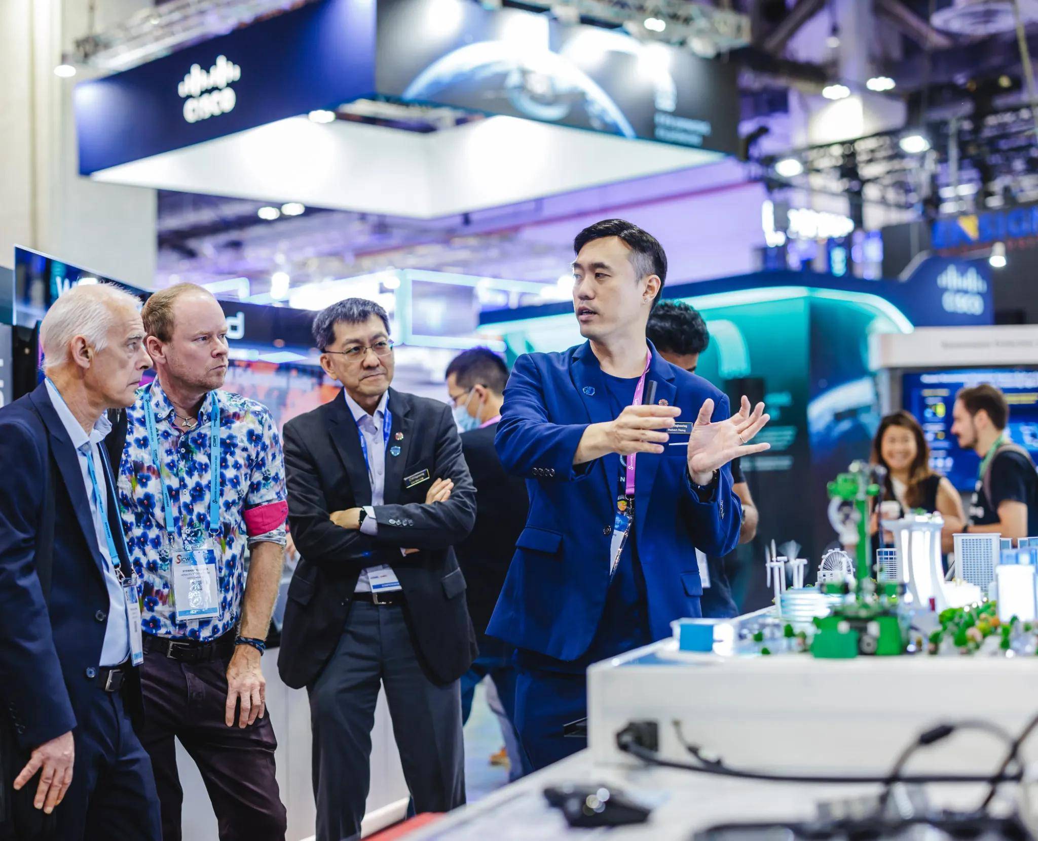 A man presents a miniature city model to three attendees at a tech exhibition booth, with a Cisco banner in the background.