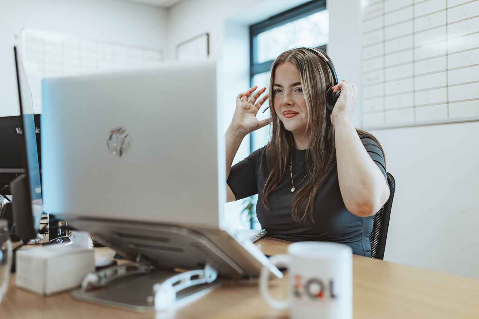 A smiling woman at a desk puts on a headset in front of a laptop.