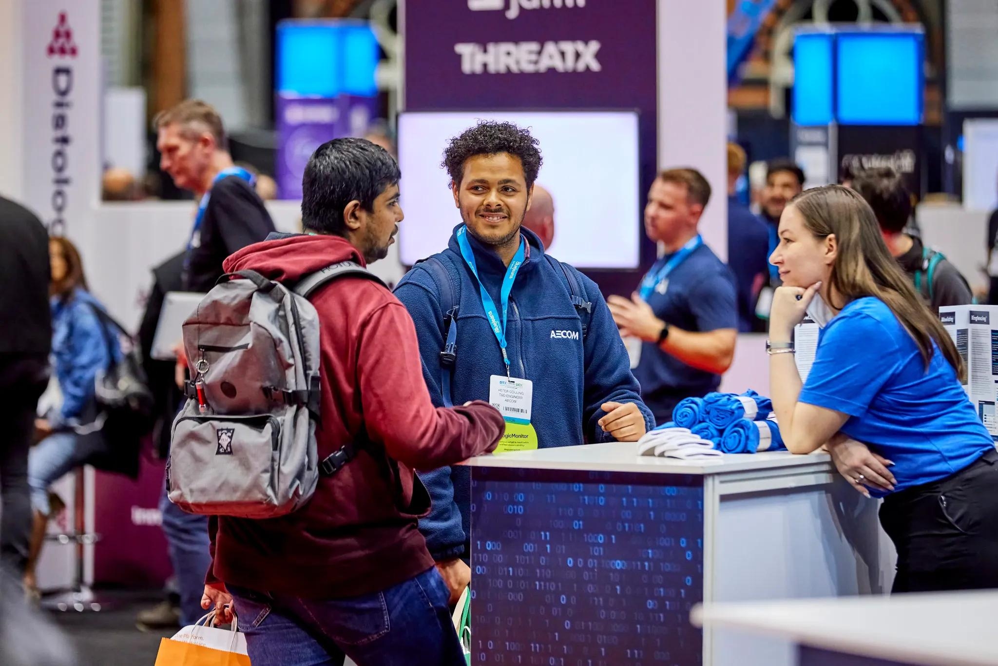 A man with a backpack talks to two smiling booth staff at a trade show.