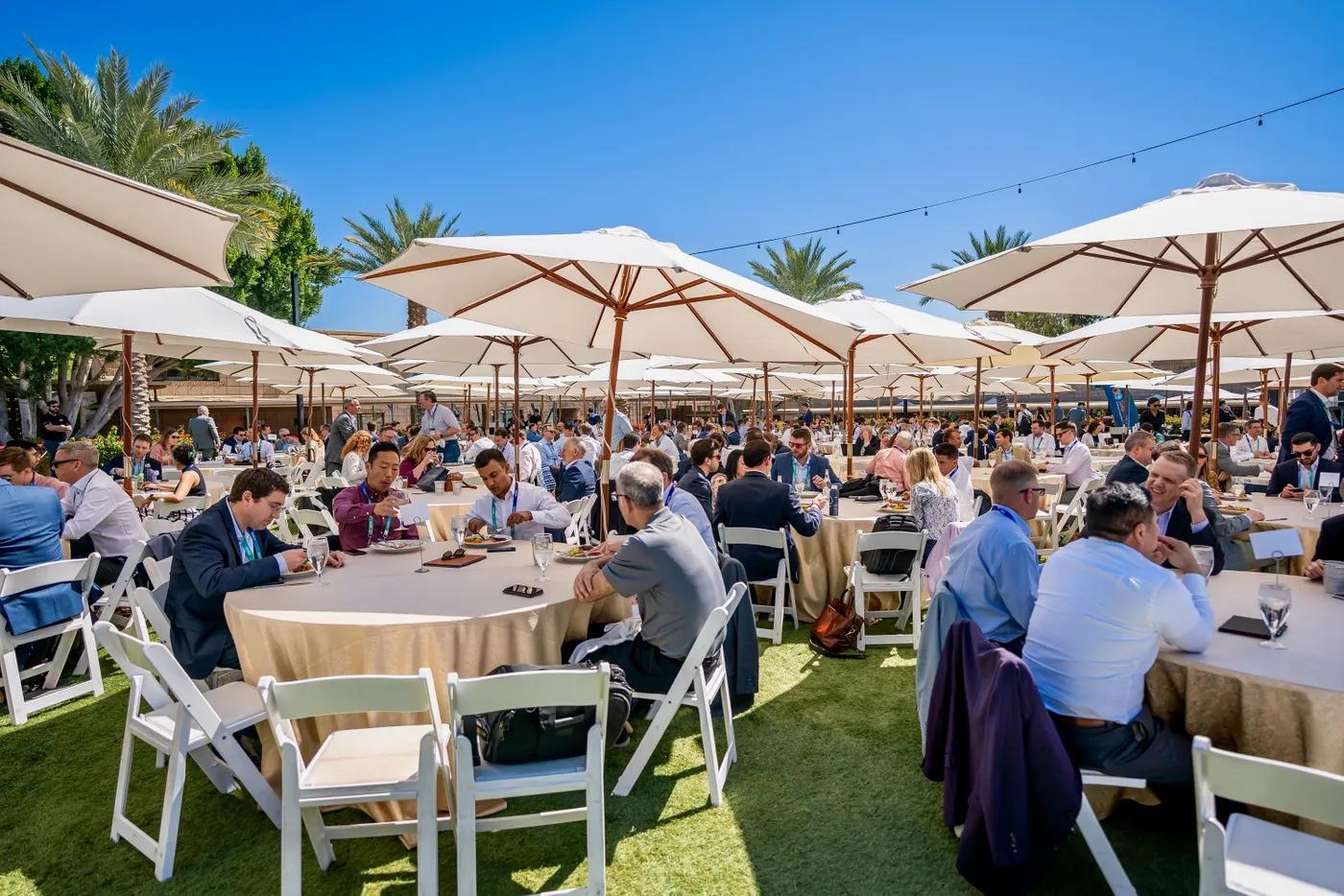 Large outdoor event with many people seated at tables under white umbrellas on a sunny lawn.