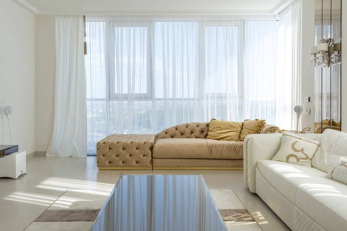 A bright living room with a tan chaise lounge, white sofa, sheer-curtained windows, and a glass coffee table.