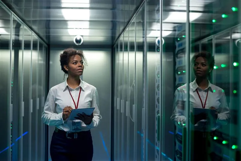 A woman with a tablet stands in a data center, her reflection visible in green-lit server racks.