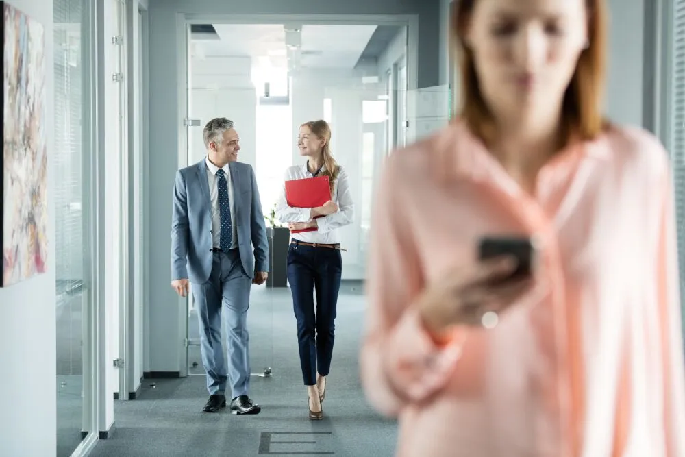A blurry woman checks her phone in the foreground, while two smiling colleagues walk and talk in a modern office hallway.