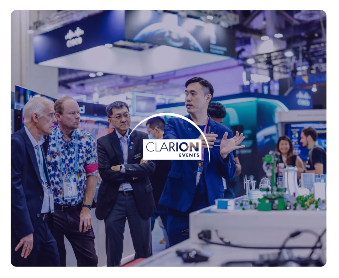 A man presents a model city at a trade show to three other men, with the Clarion Events logo overlaid.