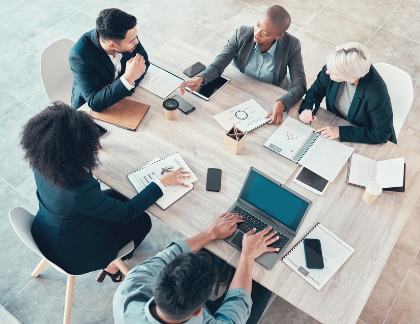 Overhead view of five diverse businesspeople discussing documents and technology at a conference table.