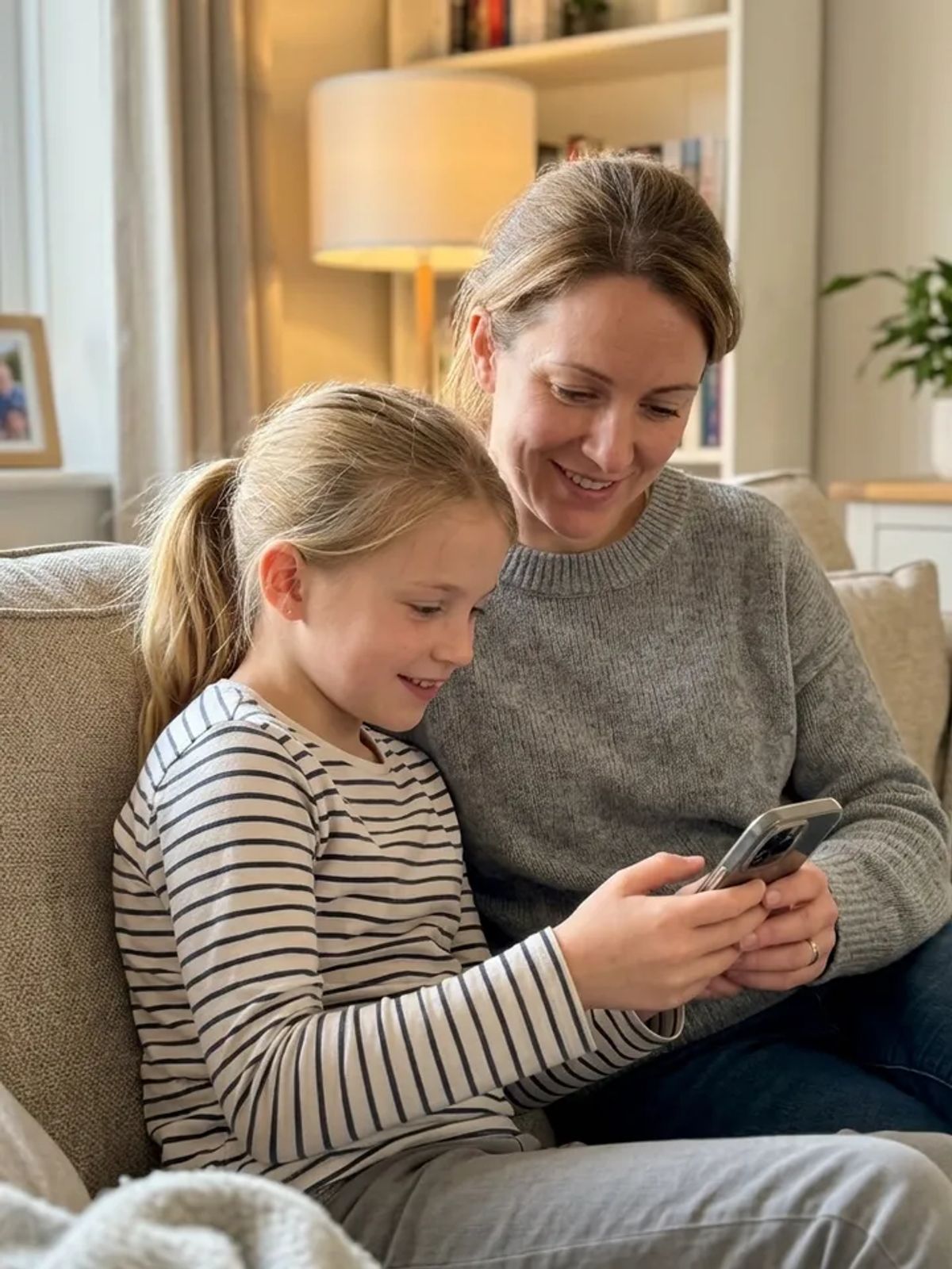A mother and her daughter sitting together on the couch, smiling as they play a game on a phone.