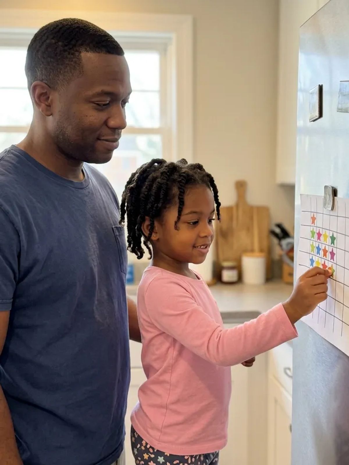 A young girl placing a star sticker on a chore chart on the fridge while her father encourages her.