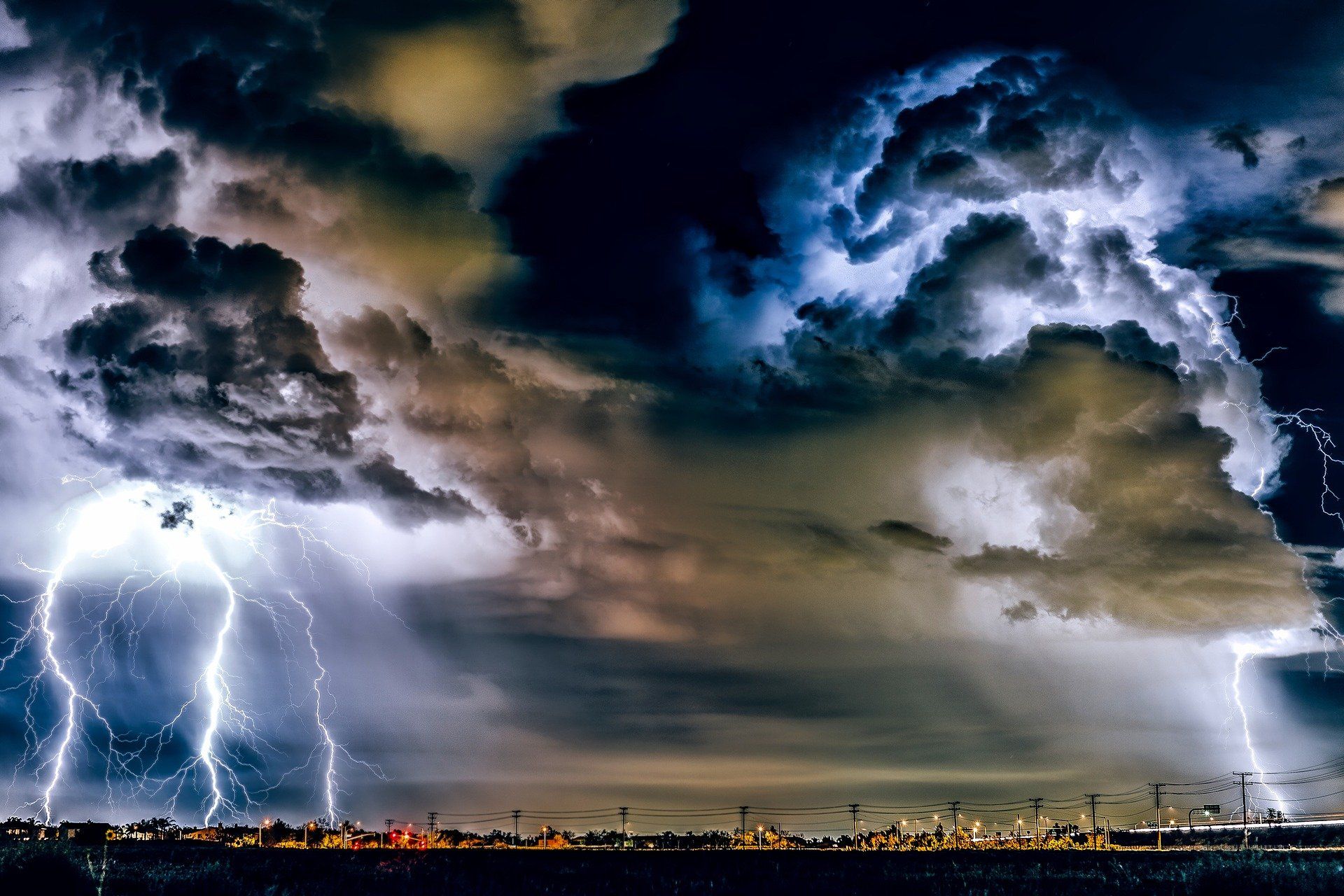 Nächtliches Gewitter mit mehreren Blitzen über einer beleuchteten Industrieanlage und Hochspannungsleitungen.