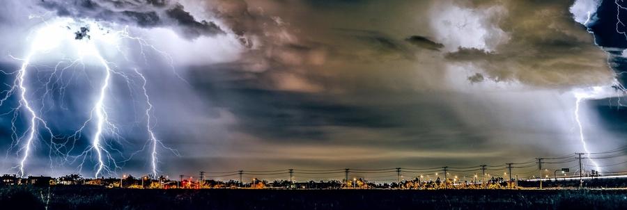 Nächtliches Gewitter mit mehreren Blitzen über einer beleuchteten Industrieanlage und Hochspannungsleitungen.
